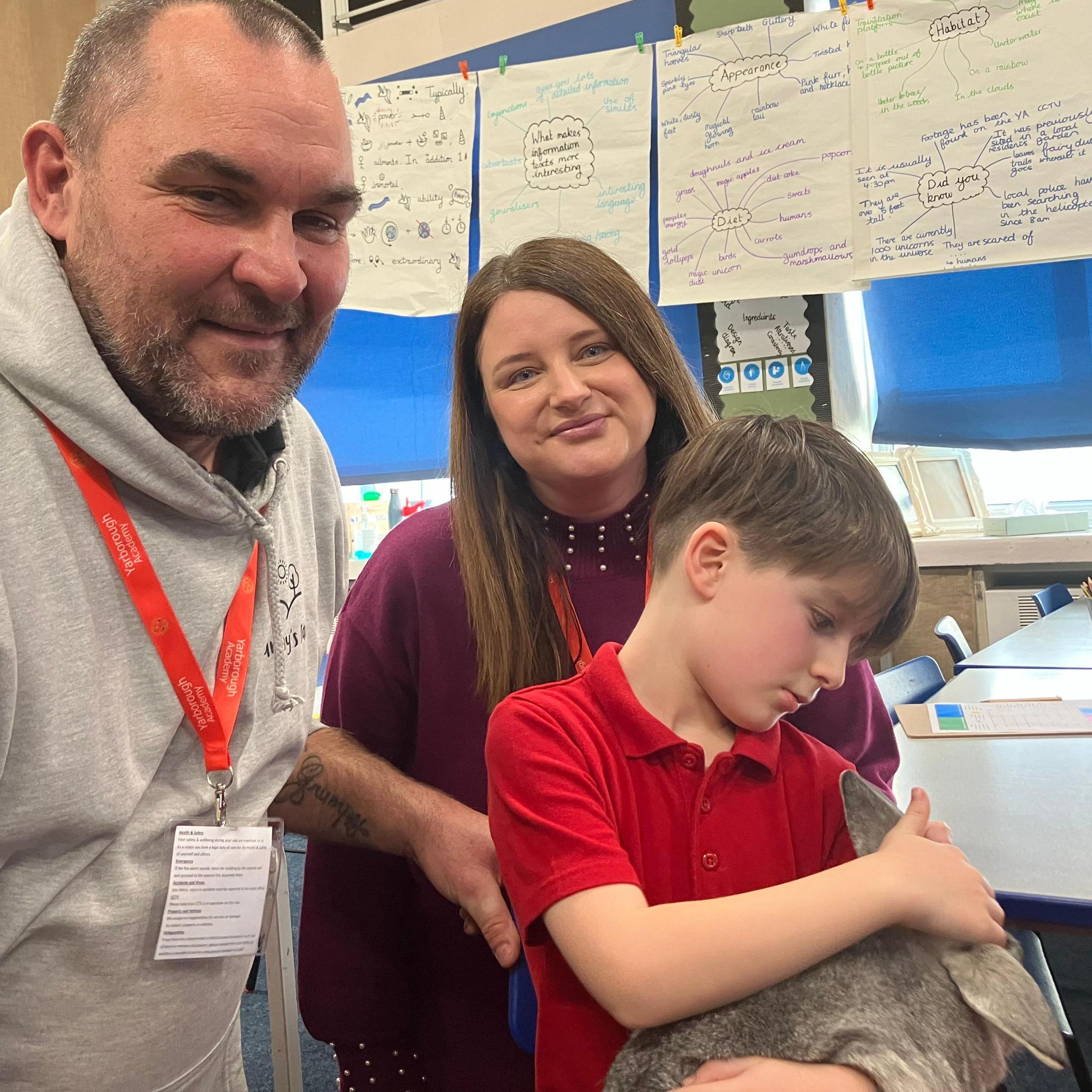 A man with a shaven head and beard smiles as he stands next to a mother and her young son, who is looking down at a grey rabbit in a school classroom. 