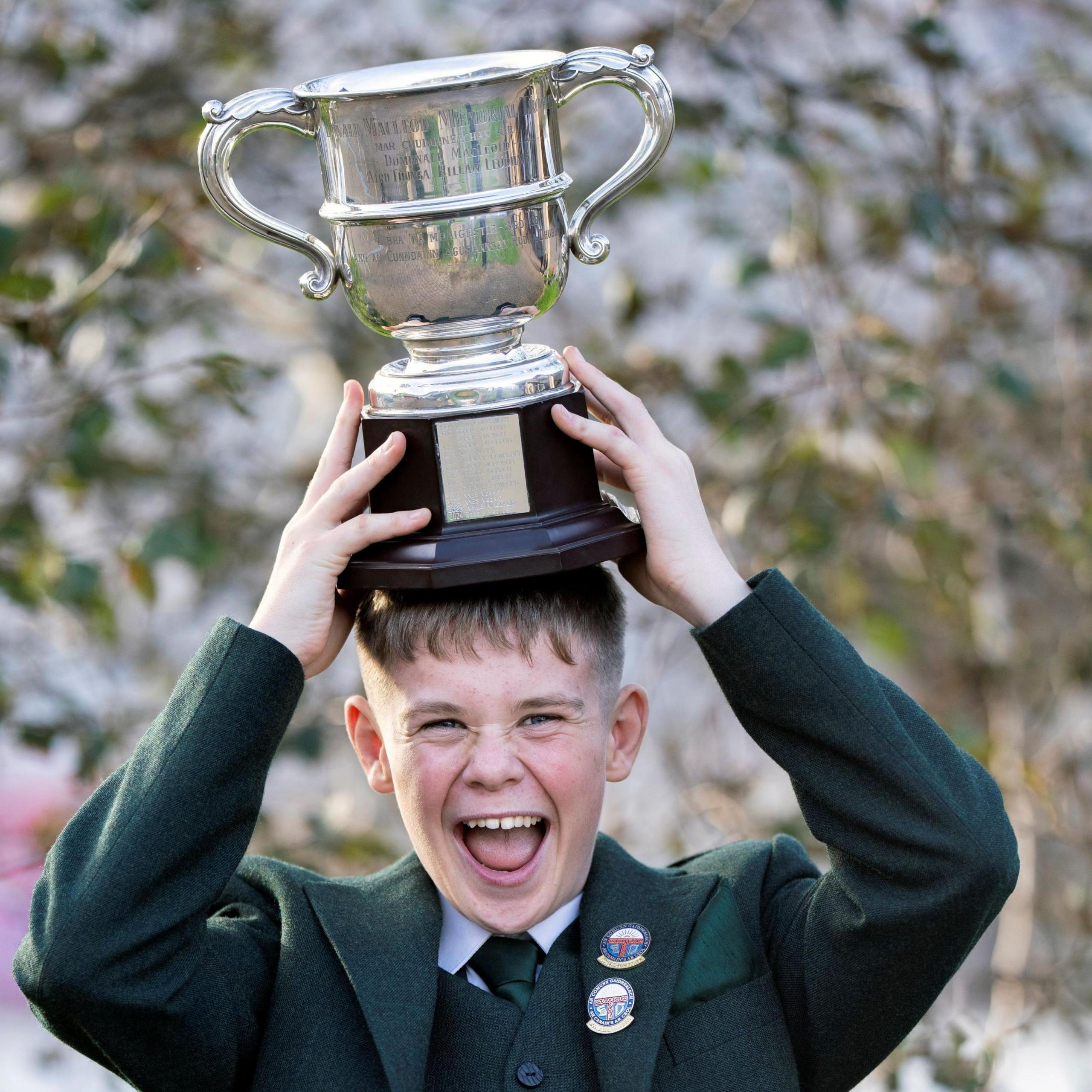 Robbie Donald MacLean is a boy with fair hair and a dark green blazer over a dark green waistcoat and white shirt with a dark green tie. He is pictured outdoors and is laughing as he holds the large trophy he won in both hands on top of his head.