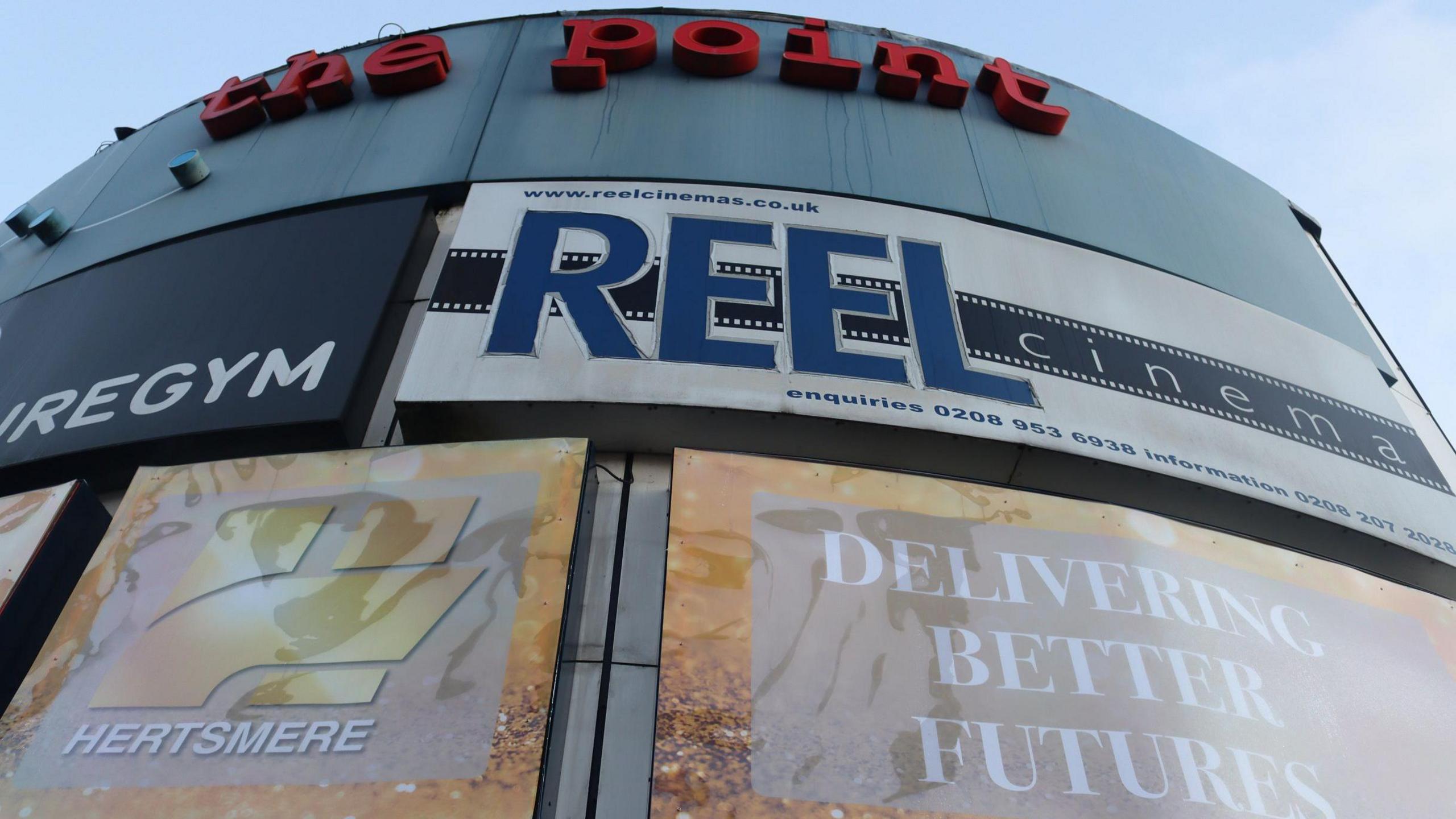 The sign for the cinema seen from outside, it is on a cylindrical building which has a red sign at the top saying "the point". Reel cinema is below in blue on a white background, and below are hoardings advertising Hertsmere Borough Council 