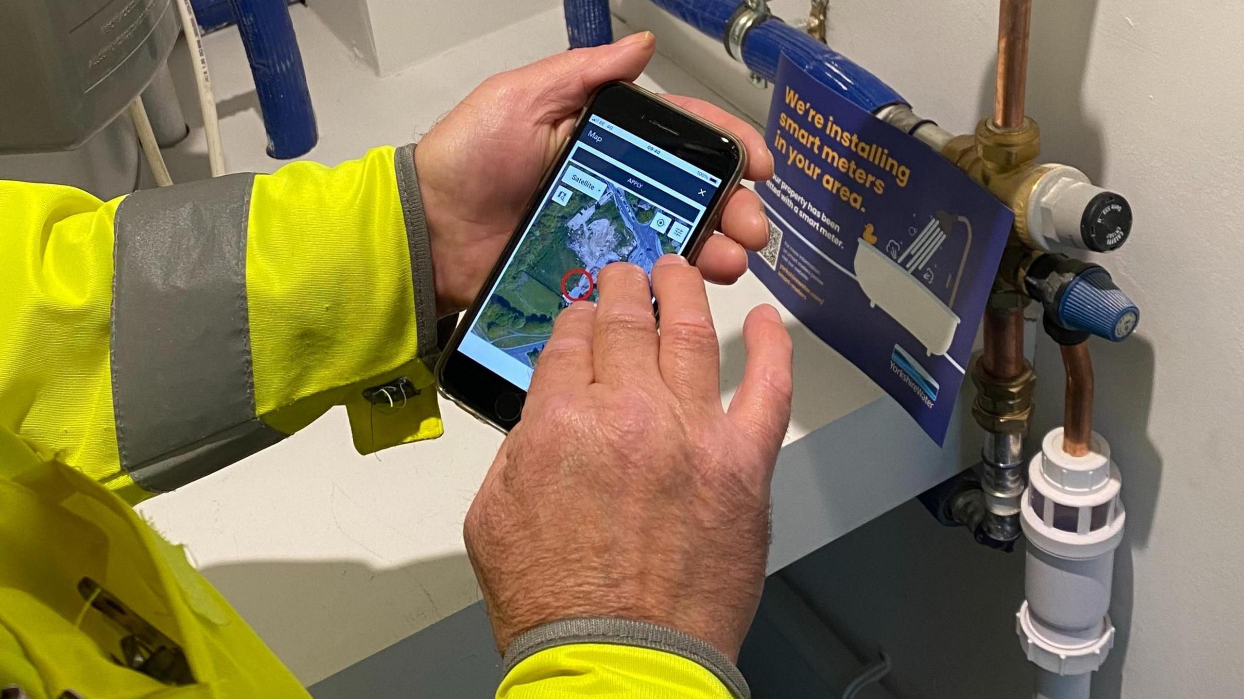 A Yorkshire Water worker in a yellow hi-vis jacket can be seen using a mobile phone to connect to a smart water meter which he has just installed.  Close-up of his hands next to an internal stop tap.