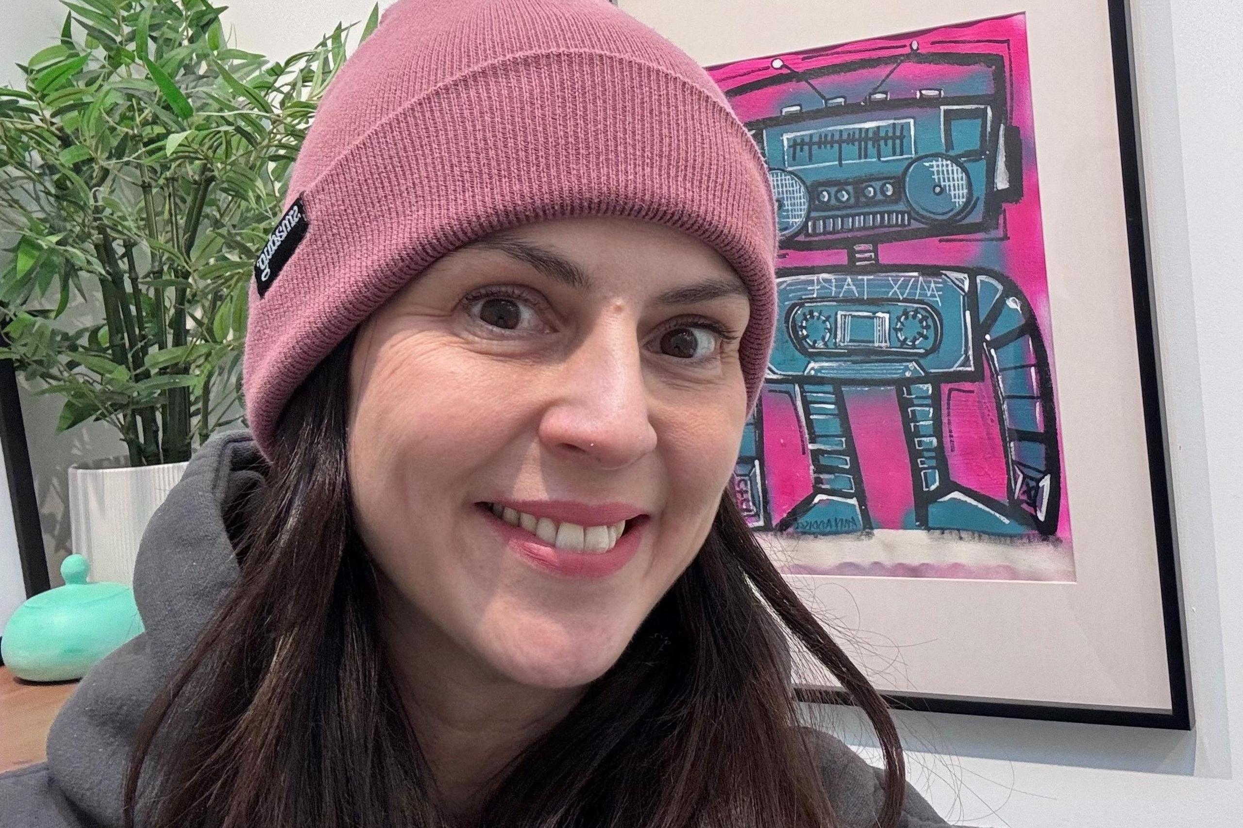 Sharon Davidson, woman wearing pink warm hat and smiling at camera, with a bright painting in the background.