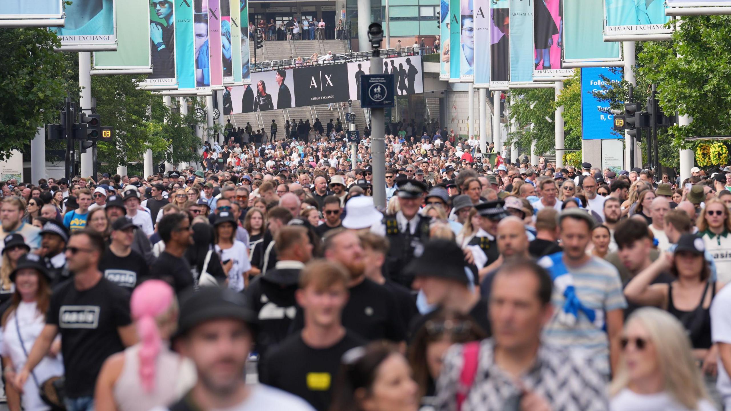 A huge crowd of Oasis fans walking down Wembley Way ahead of a previous gig. Many are wearing black bucket hats. Colourful banners line the wide walkway. In the background, grey stairs lead to the railway station.