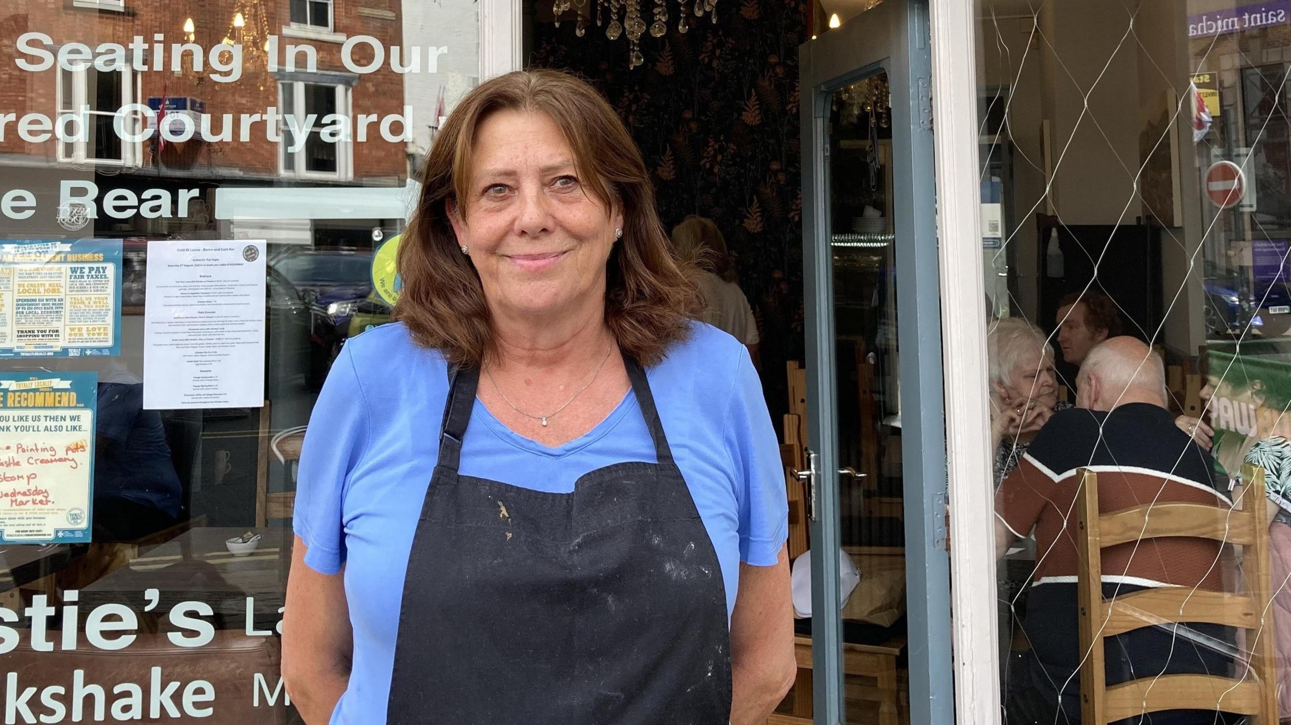 Jules Green, owner of Café Di Lucca stands outside her business. She has medium length brown hair, and wears a blue top with a black apron over the top. Customers can be seen through the café window sitting at tables.