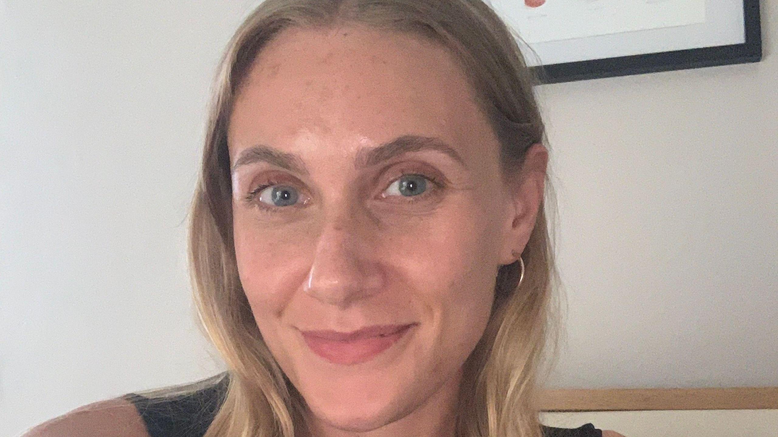 A cropped pictures of Lili Vachon, who has blue eyes and strawberry blonde hair, smiling in front of a white wall with a picture frame seen behind her head.