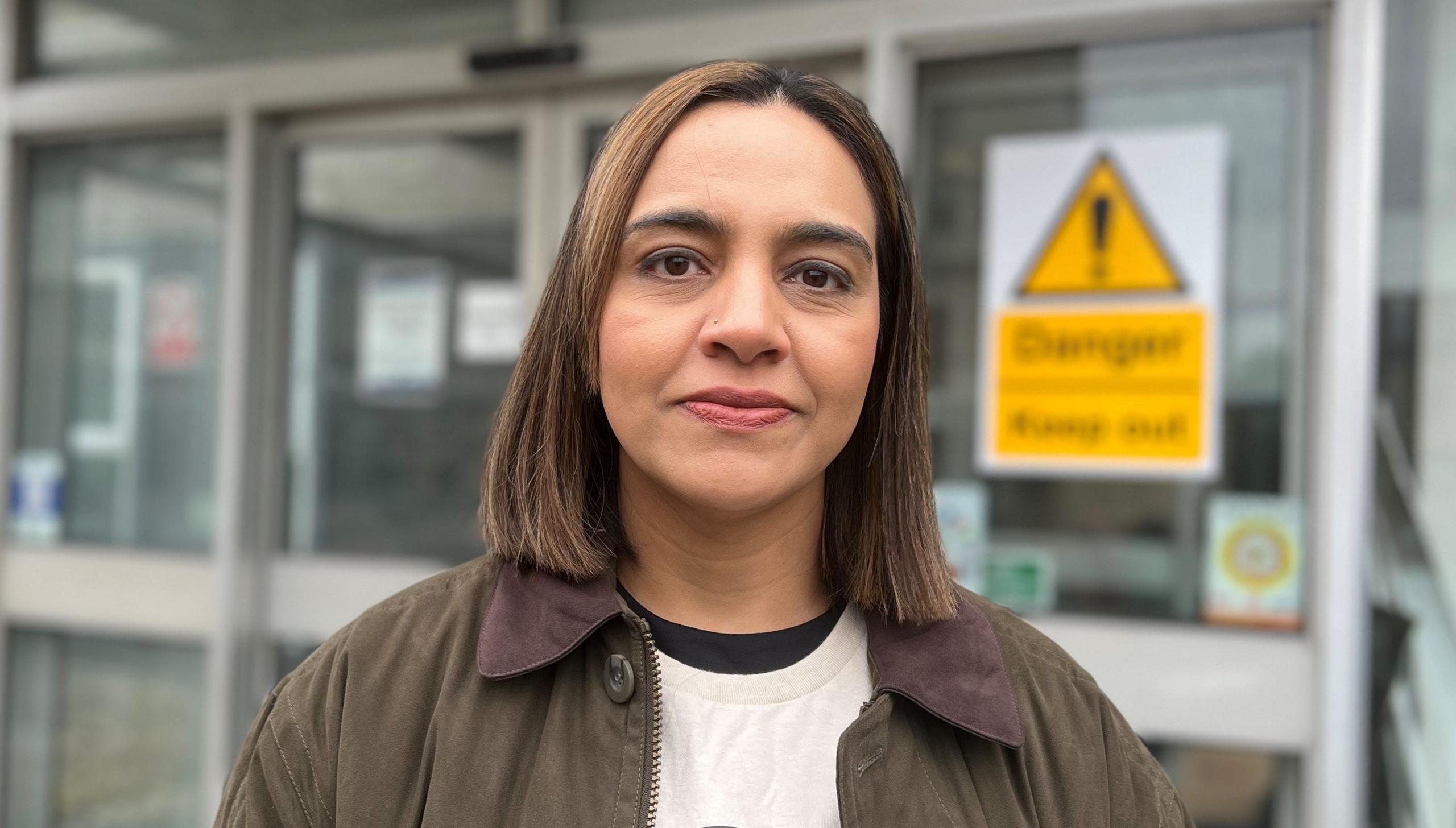 A woman with short brown hair and brown eyes standing outside some glass entrance doors. The woman is wearing a white t-shirt with a black top underneath and a brown jacket. On the door behind her is a yellow warning notice with an exclamation mark but the text is blurred so that it is not visible.