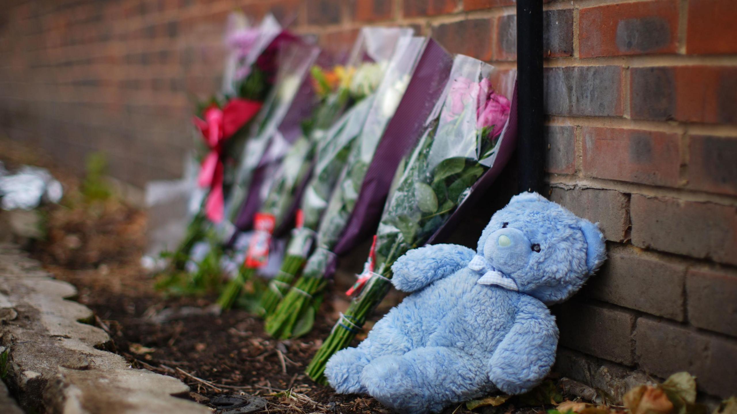 A blue teddy bear and bunches of flowers rested against a brick wall at the scene of the fire