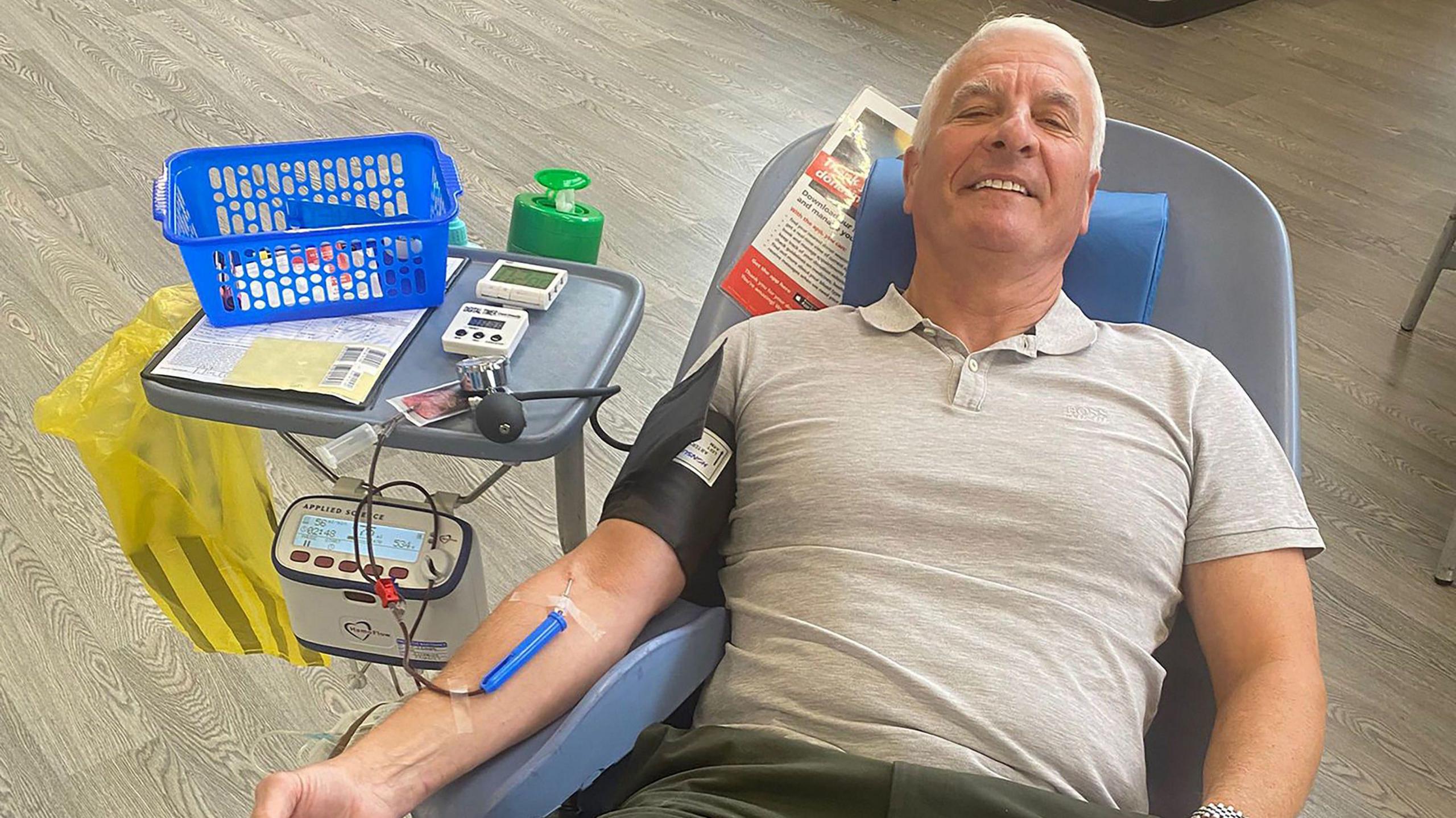 A man with short white hair smiles at the camera as he lies back in a medical chair with a needle in his arm connected to a tube carrying blood to a collection device. He is wearing a grey polo shirt and has a blood pressure monitor on his arm. A small table connected to the side of the chair has medical devices and a blue plastic basket on it. 