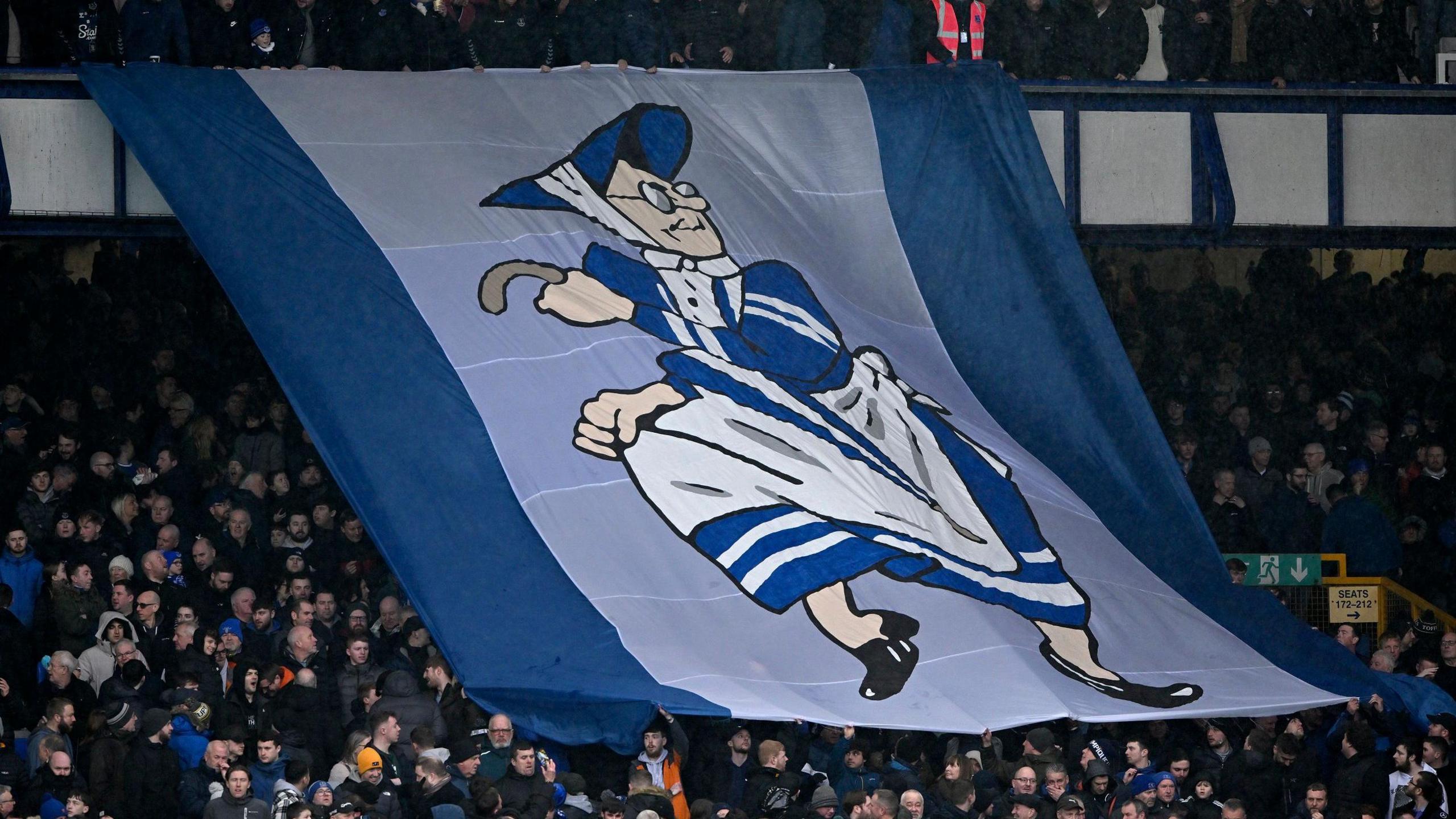 Large flag at Goodison Park being passed over the heads of fans. It features a cartoon image of an old woman dressed in the Everton toffee lady uniform of a blue dress and white pinny.