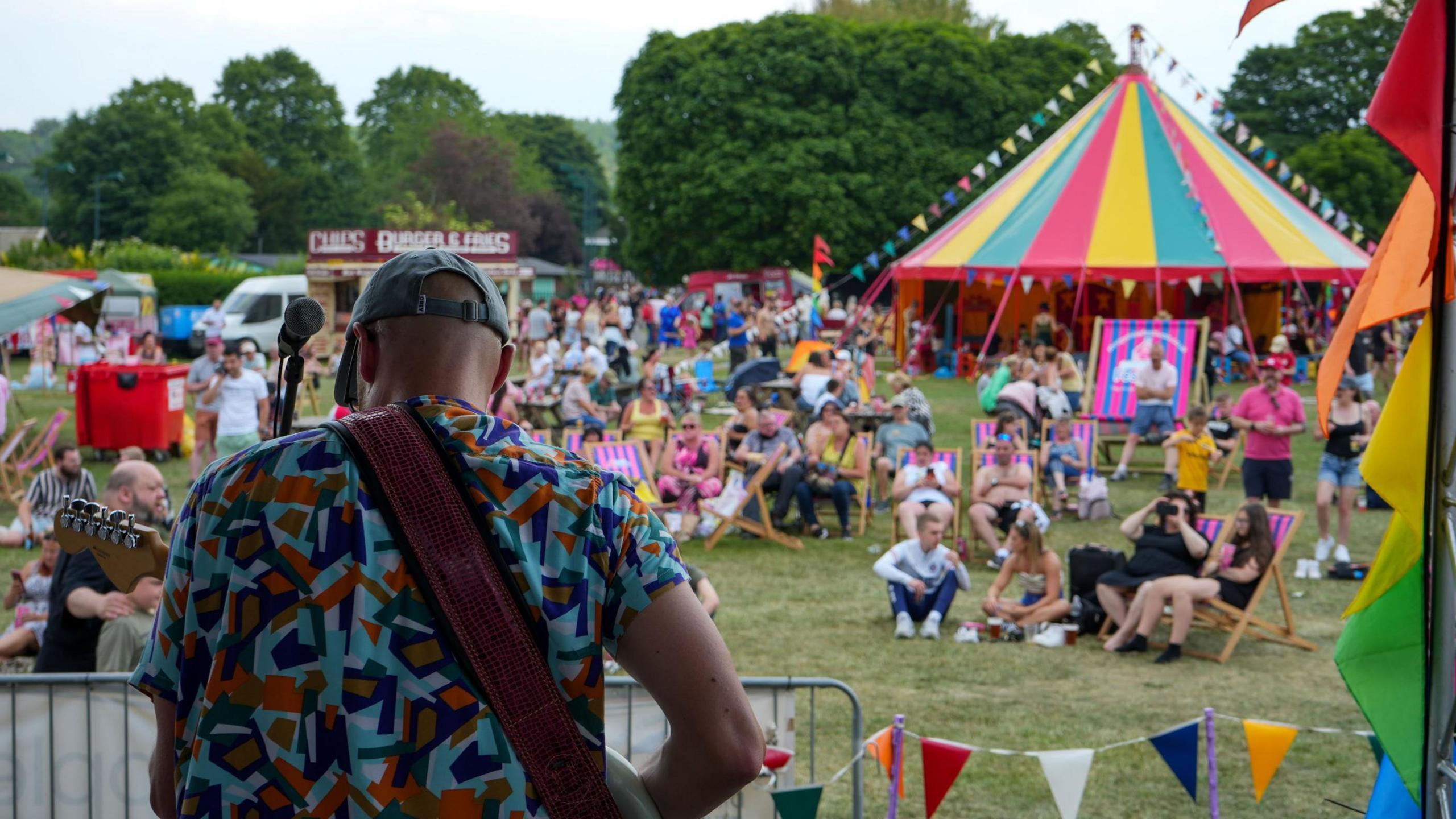 A photo from the 2023 event which shows the back of a man who is playing a guitar and wearing a baseball cap and patterned shirt which had colourful shapes on it. In front of him, people are sitting on the grass or on deckchairs which are pink and blue striped. There is a colourful teepee style tent in the background. 