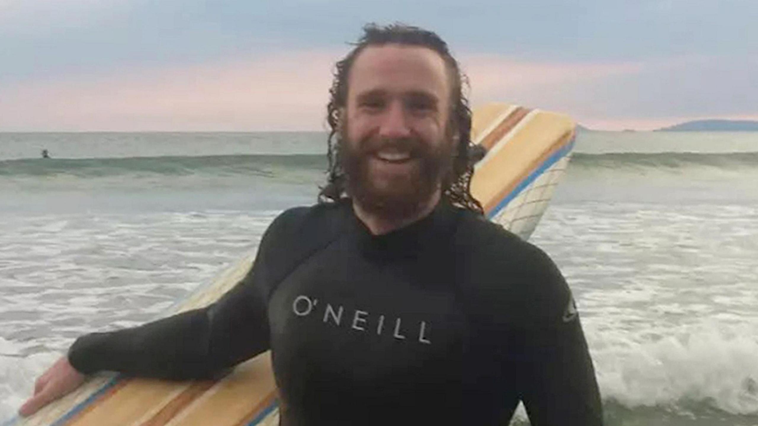 Alexander Barrett wearing a wet suit and holding a surf board while standing in front of the sea. He is smiling and has long brown hair and a brown beard.