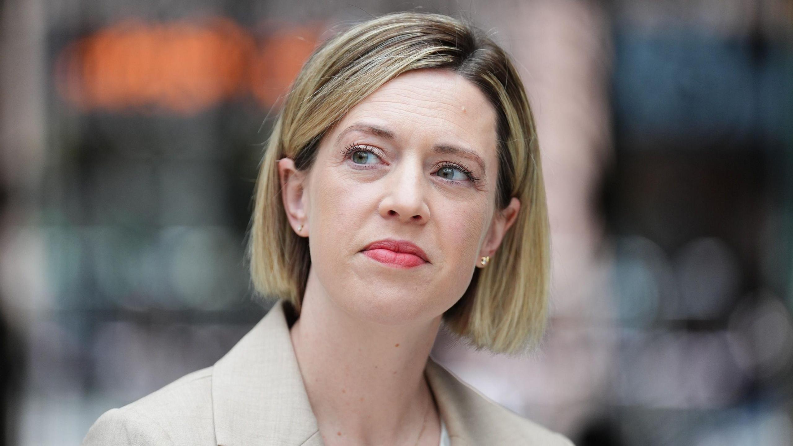 A woman with a blonde bob, wearing a beige blazer and a white top staring off into the distance.