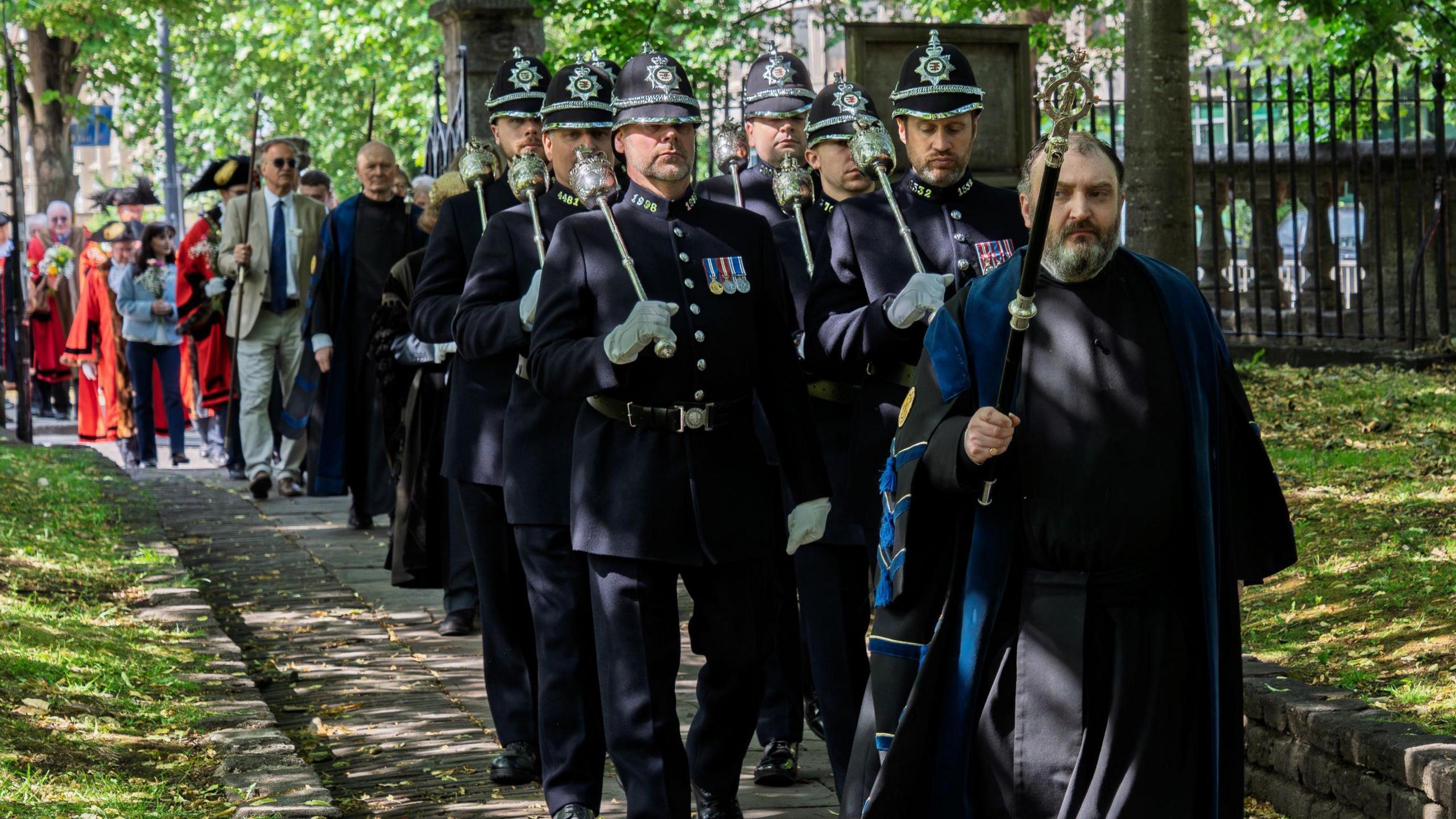 A man carrying a wooden staff and dressed in black clothes leads a line of people dressed in traditional early 20th Century police uniforms into the churchyard of St Mary Redcliffe in Bristol as part of its annual Rush Service.