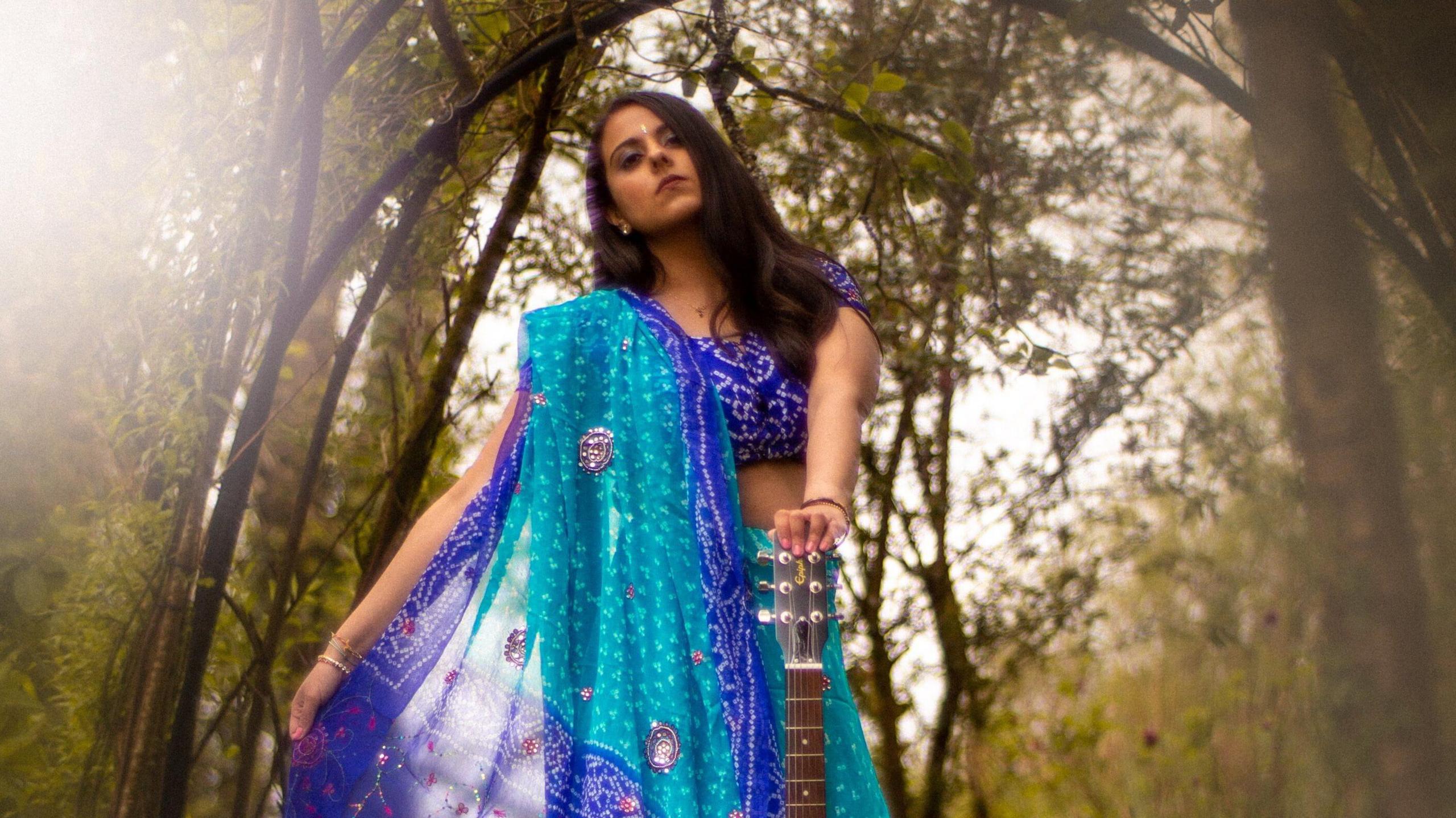 Maya Lakhani  in a forest wearing a sari and holding a guitar