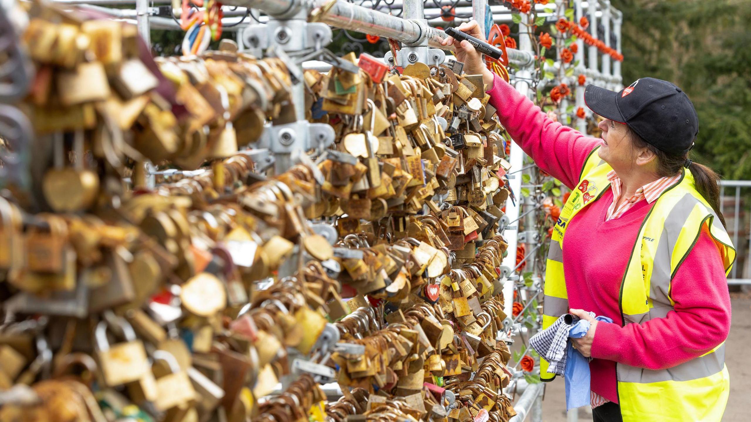 Rows of padlocks on a structure with a woman on the right - Emma Harrison - wearing a hi-vis jacket looking at it, with her hand on it.