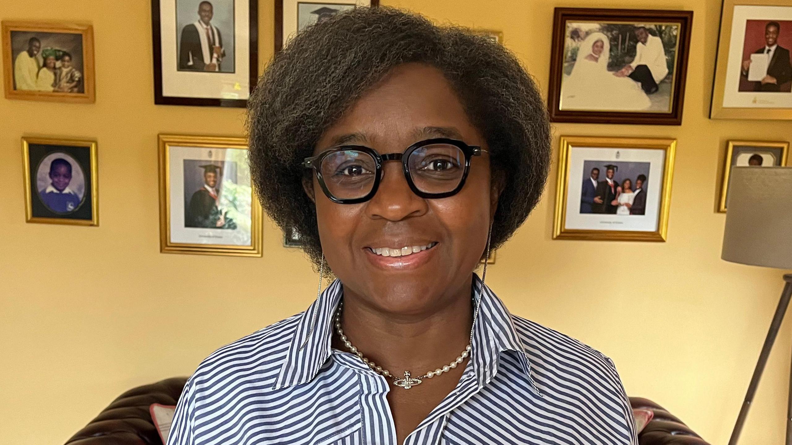 Linda Nnene sitting in front of a yellow painted wall with framed photographs on it. She is wearing a blue and white striped shirt, a necklace and black framed glasses.