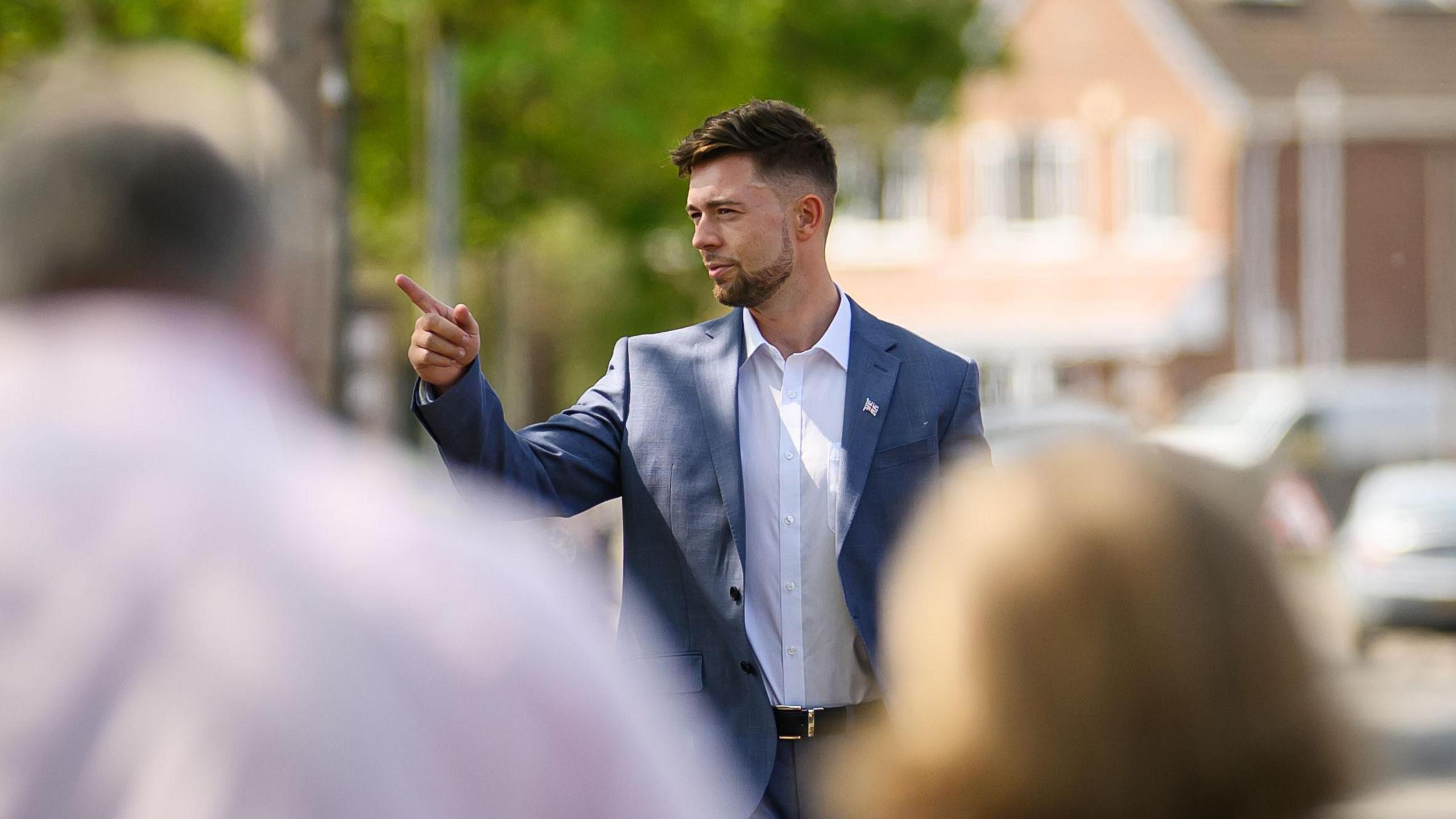 A man in his thirties wearing a blue suit jacket and white shirt. He is stood in a public street and pointing to something.
