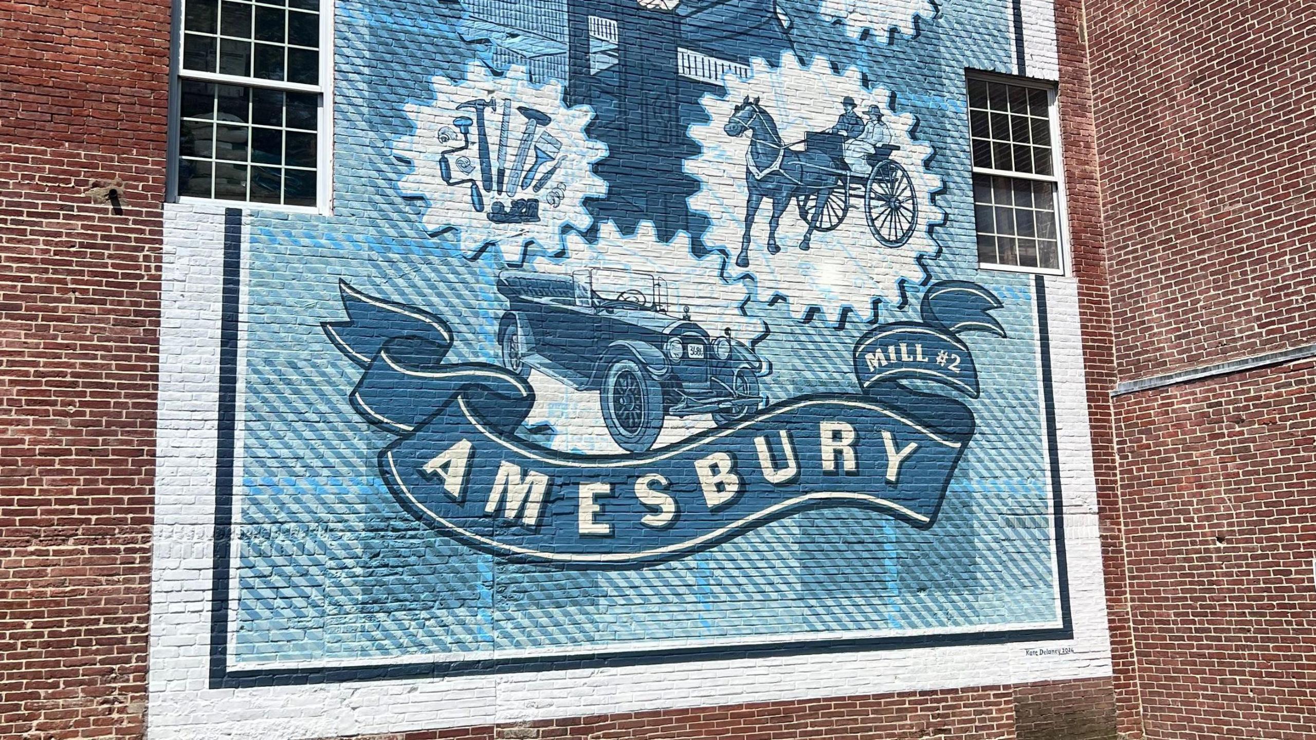 A mural on a red brick wall. The blue and white mural, in the style of a postage stamp has images of an early motor car, a selection of tools and a horse and cart carrying two people. Below the images is a banner which reads "Amesbury" in capital letters and "Mill #2". 