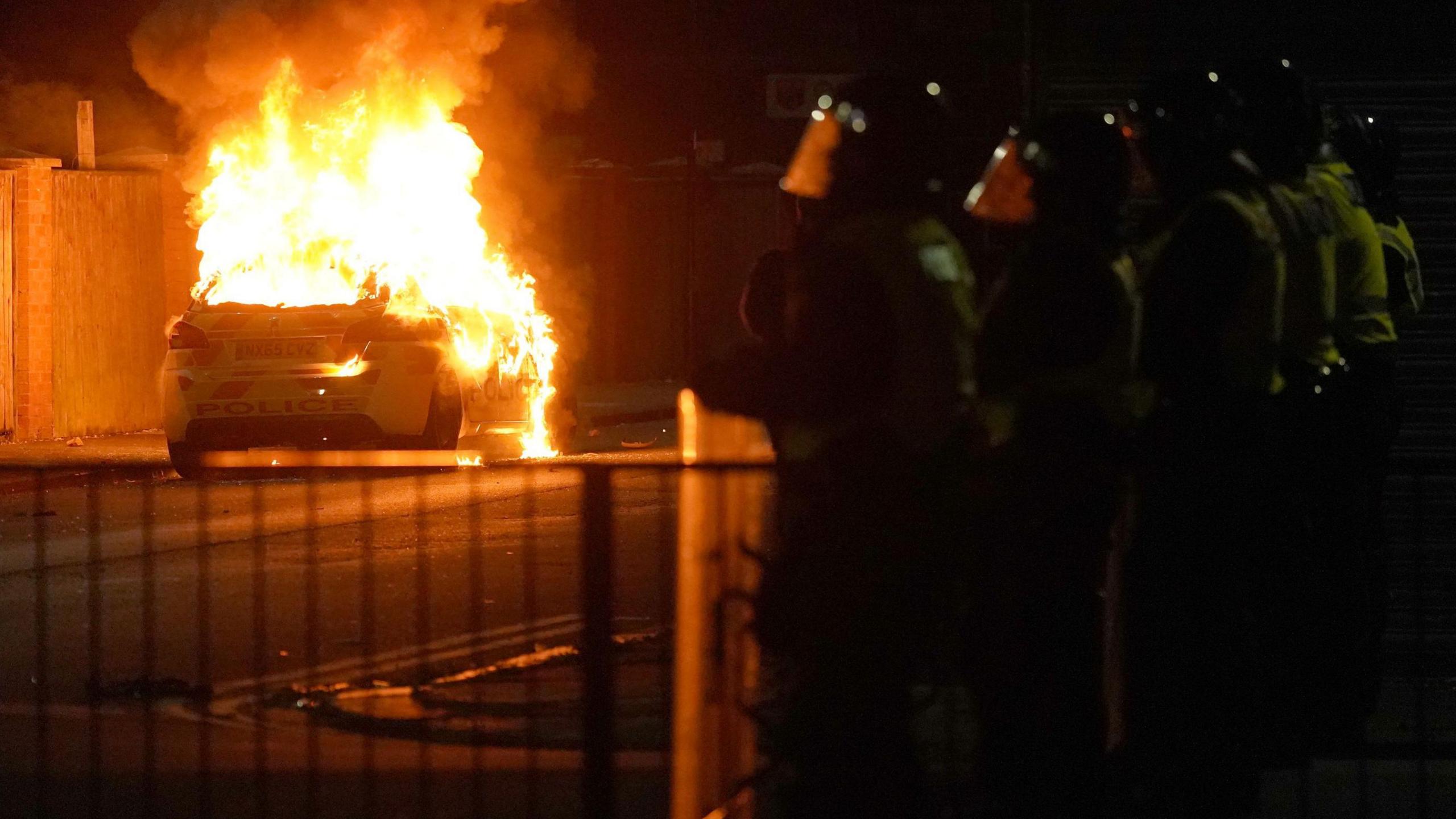 A police car is on fire. Policemen watch and film from the sidelines