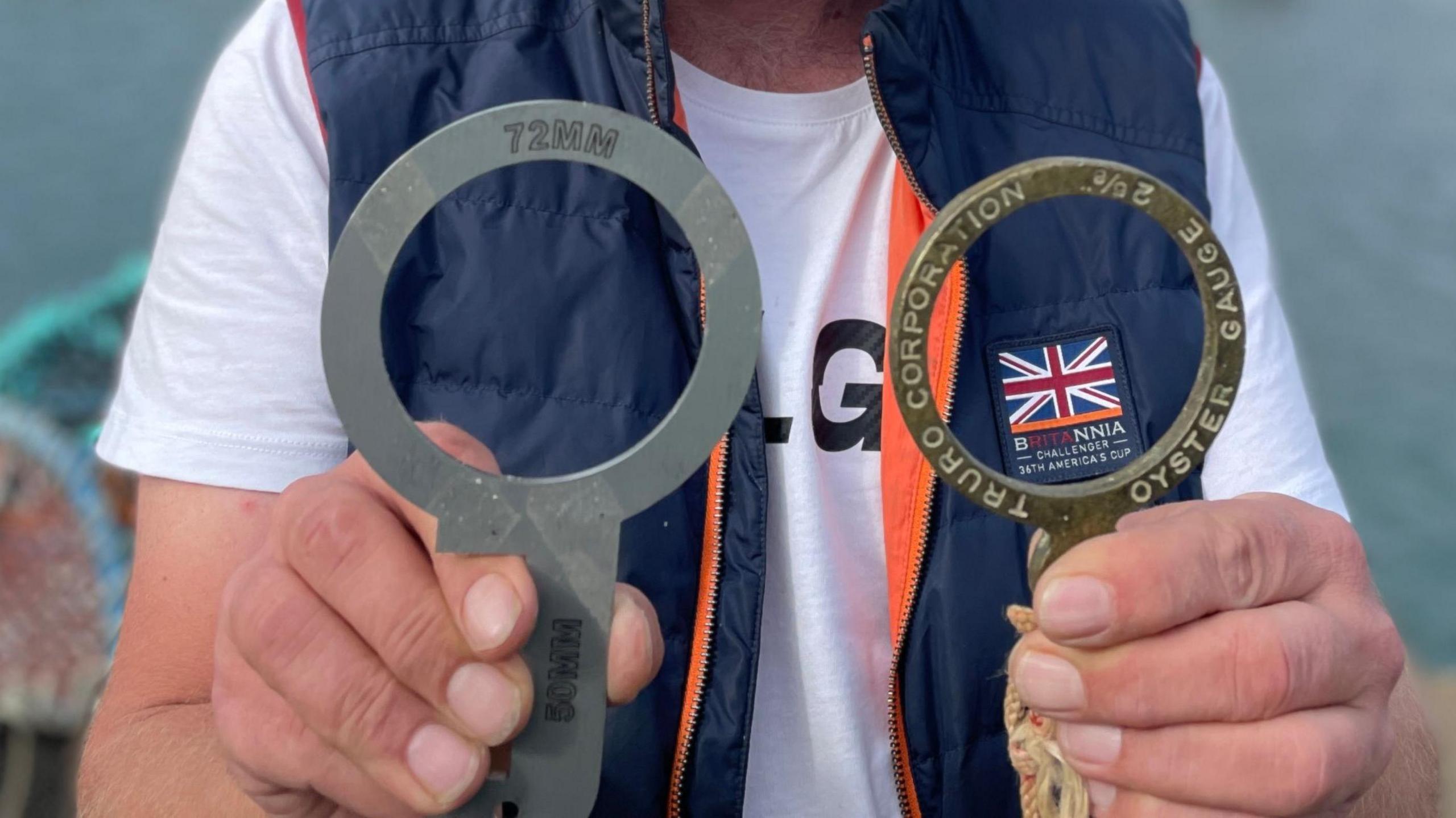 A close of a man's hands holding two metal rings which are used for measuring the size of oysters. One is silver-coloured and is larger than the other brass one. The brass one has engraved around it: TRURO CORPORATION OSYTER GAUGE 2 5/8 while the other one reads 72MM.