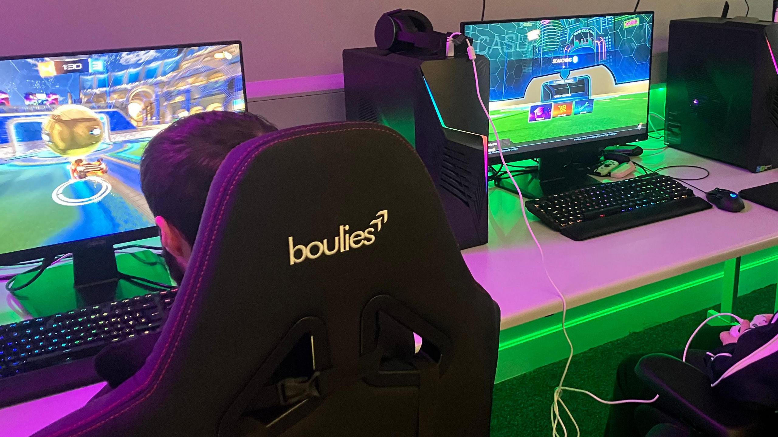 Two boys sit on black gaming chairs in front of computer monitors playing different games. The room is lit with pink and green lights.