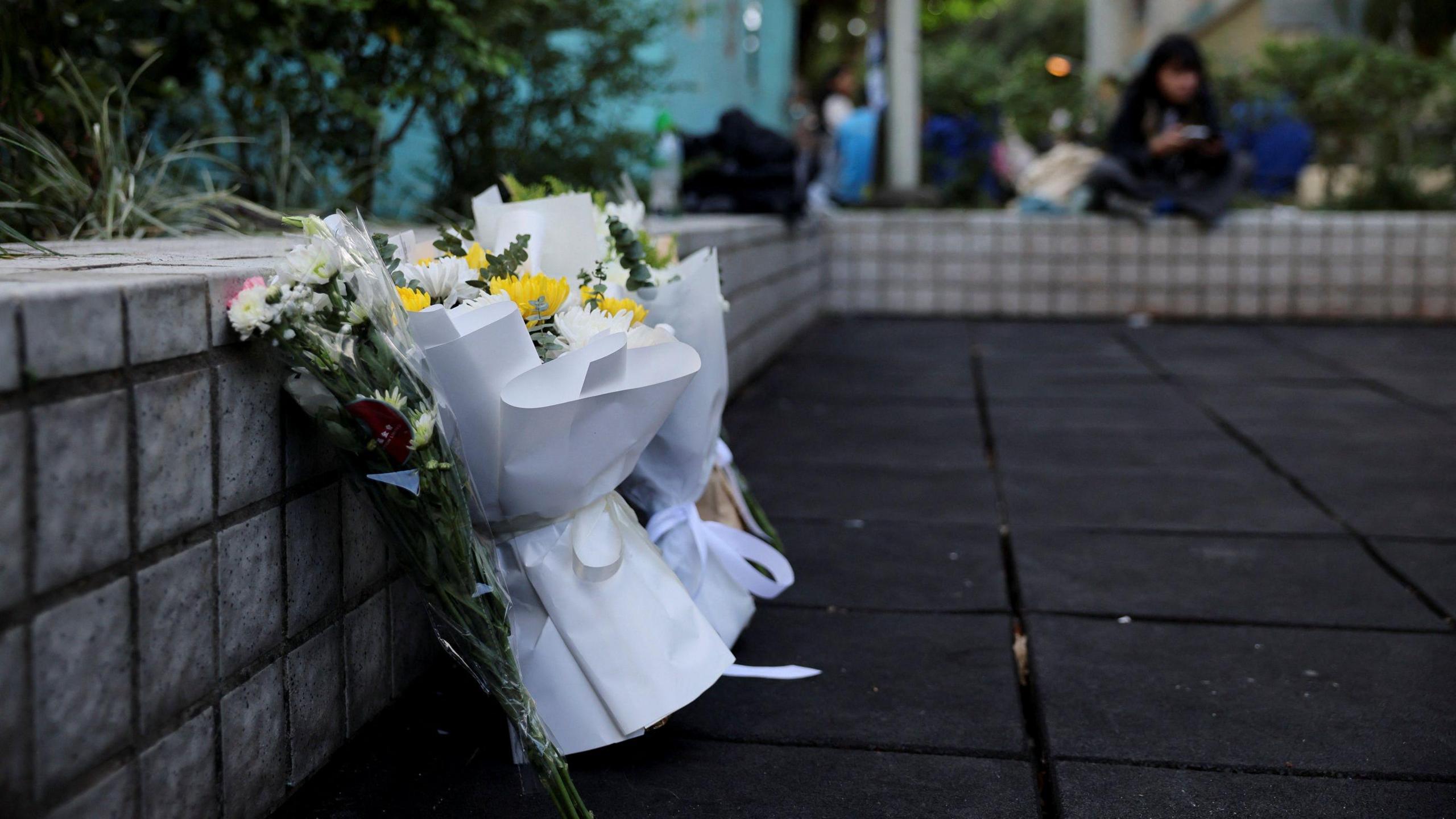 Three flower bouquets leaning against a ledge on the ground outdoors. There are people sitting on the ground