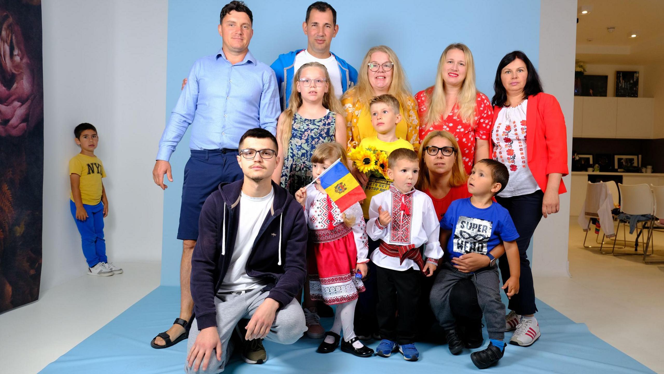 A Romanian group, posing for a photo, in a studio, one child is holding a flag, while another is away from the camera standing in a corner