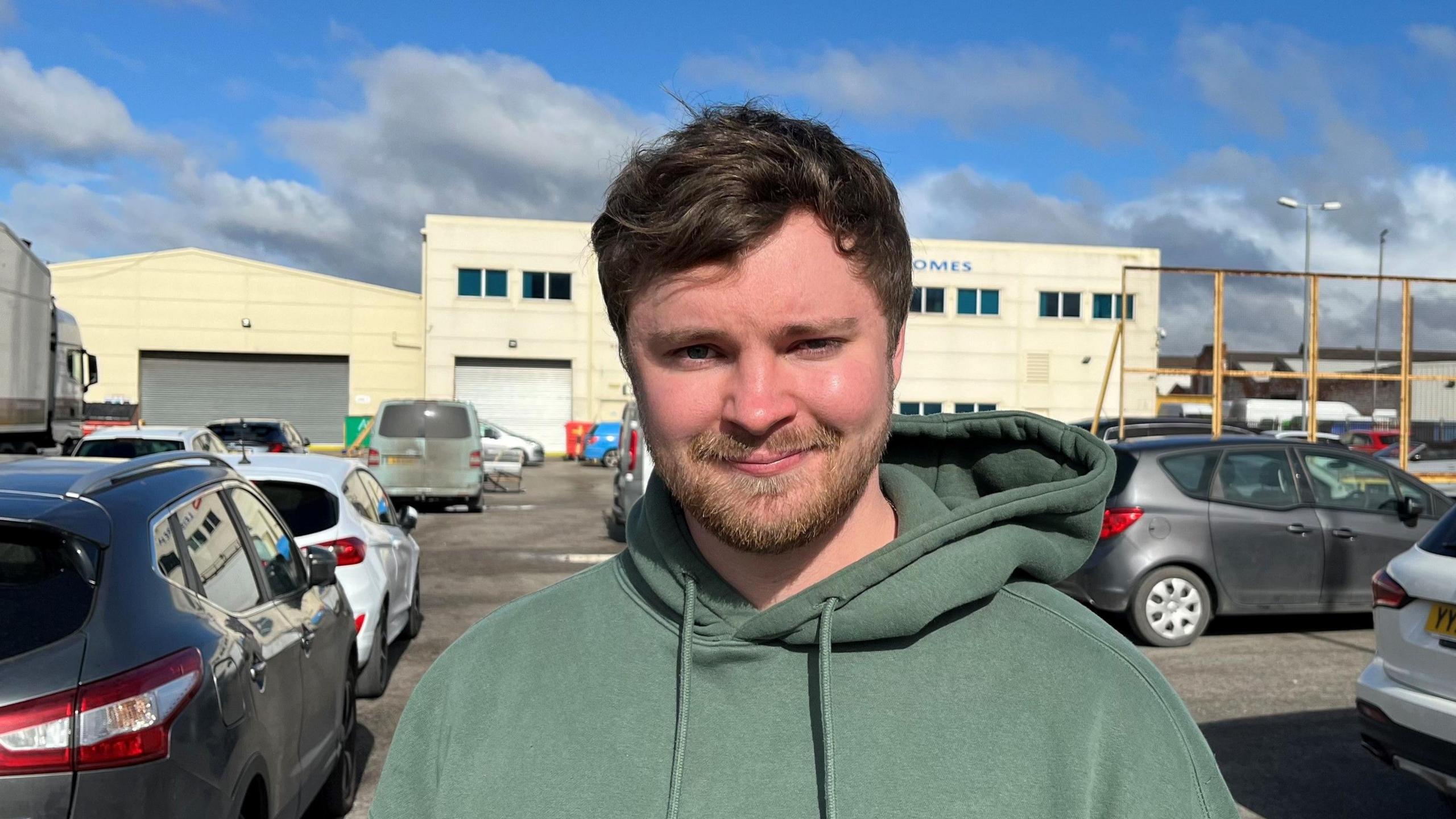 Jack McLaren standing outside the Atlas Leisure Homes headquarters on Wiltshire Road, Hull. He has dark brown hair and a beard and is wearing a green hoodie. Behind him is a large, cream-coloured building surrounded by cars.