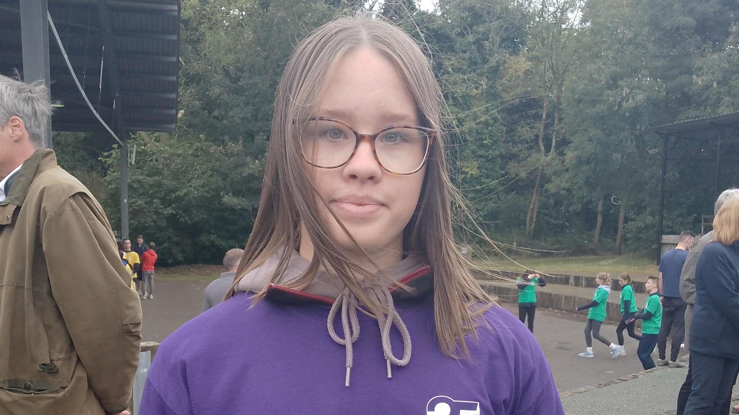 Rebecca Darling is wearing a purple hoodie. She wears tortoiseshell glasses and has brown hair. There are young people wearing green in the background.