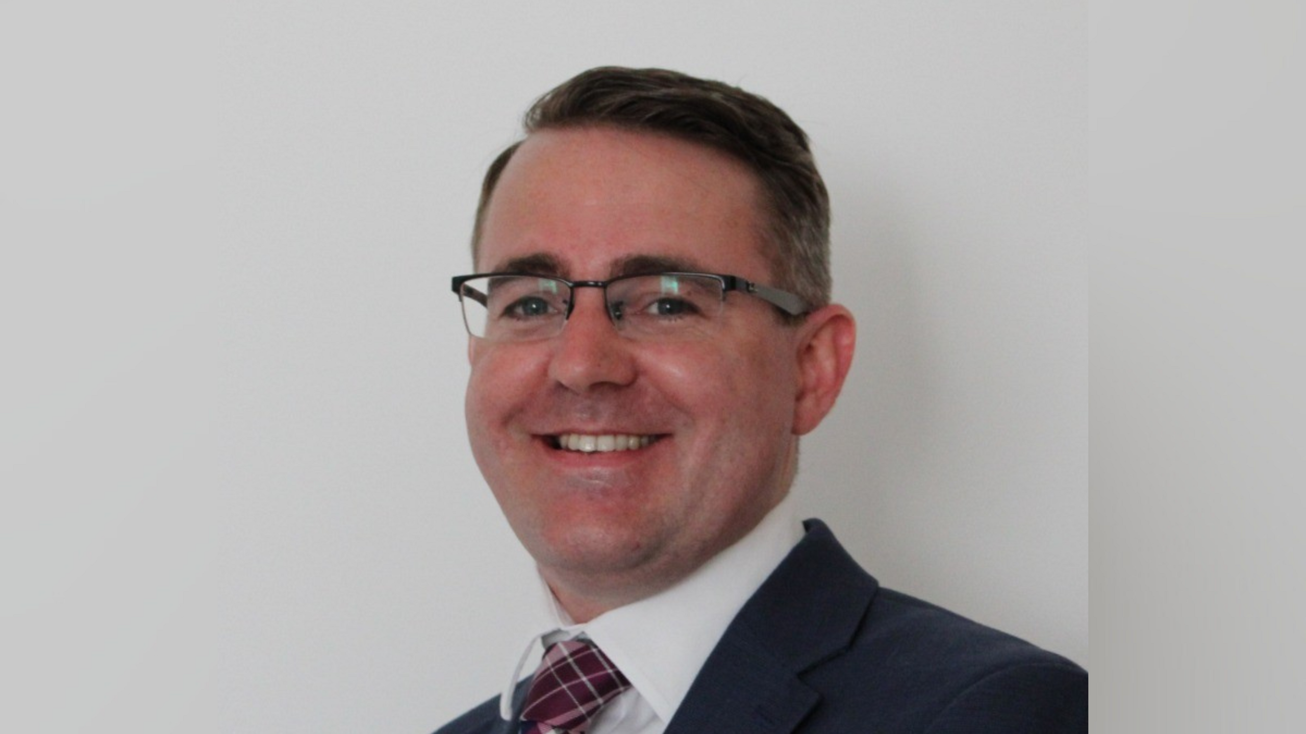 A head and shoulders image of Rory Chamberlain. He is smiling and wearing glasses, a tie, a white shirt, 