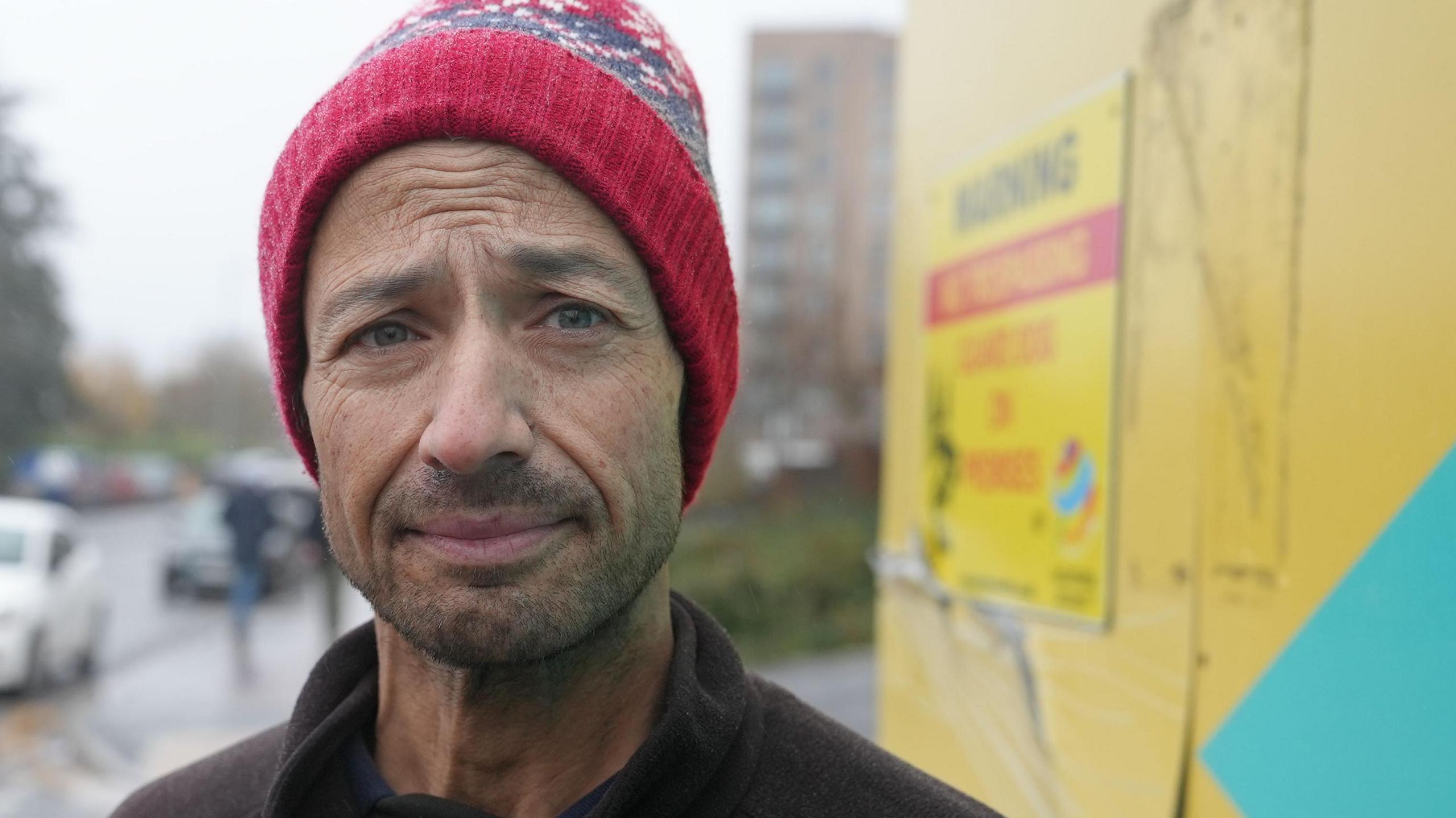 Andy wearing a red bobble hat and black jumper. He is looking at the camera without much expression on his face. He is stood next to yellow boarding and has a car going past him to the left of the image.