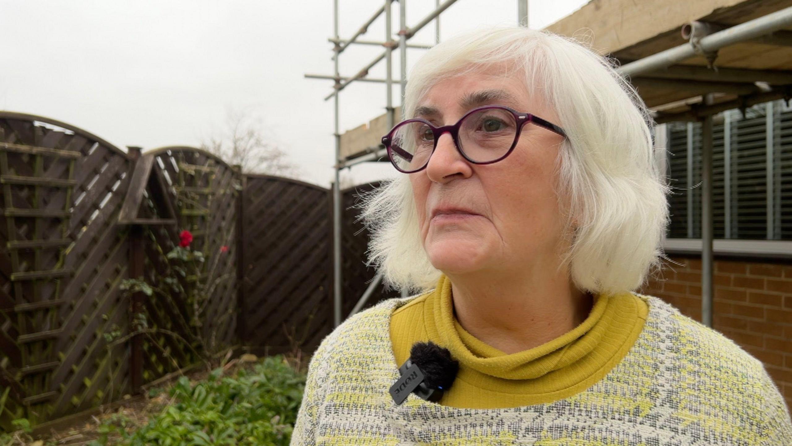 A woman in a checked yellow jumper and yellow polo neck stands in front of a bungalow, which has scaffolding on the front.