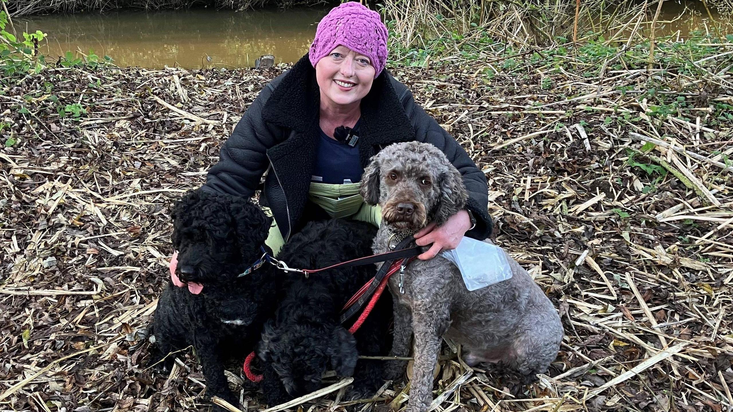 A woman wearing a pink crochet hat, black fur lined jacket and yellow trousers crouches on the ground with her arm around three medium sized dogs on leads. Two of the dogs are black and the third, on the right, is a grey to brown colour. Behind them is a river.