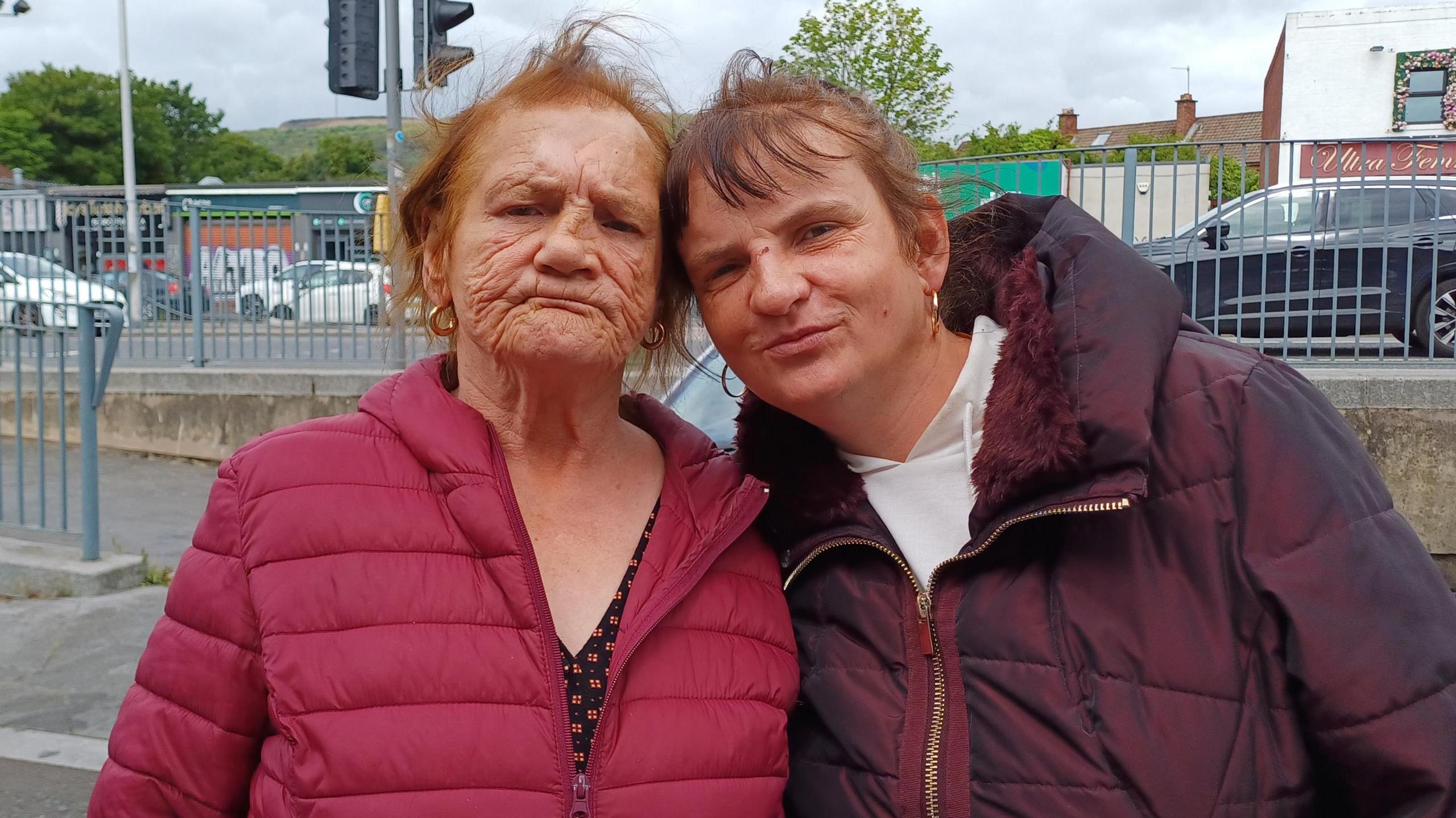 Two women are standing in the street with their heads against each other. One is wearing a red puffy coat and the other is wearing a maroon coat. They are both wearing gold hooped earrings.