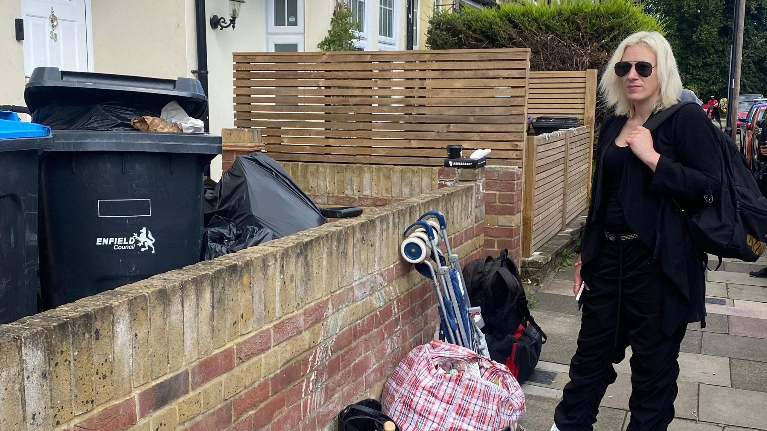 Carly, wearing black sunglasses and a black top, jacket and trousers and white trainers, stands outside a wall of the house she lived in. On the side of the wall are rubbish bins. 