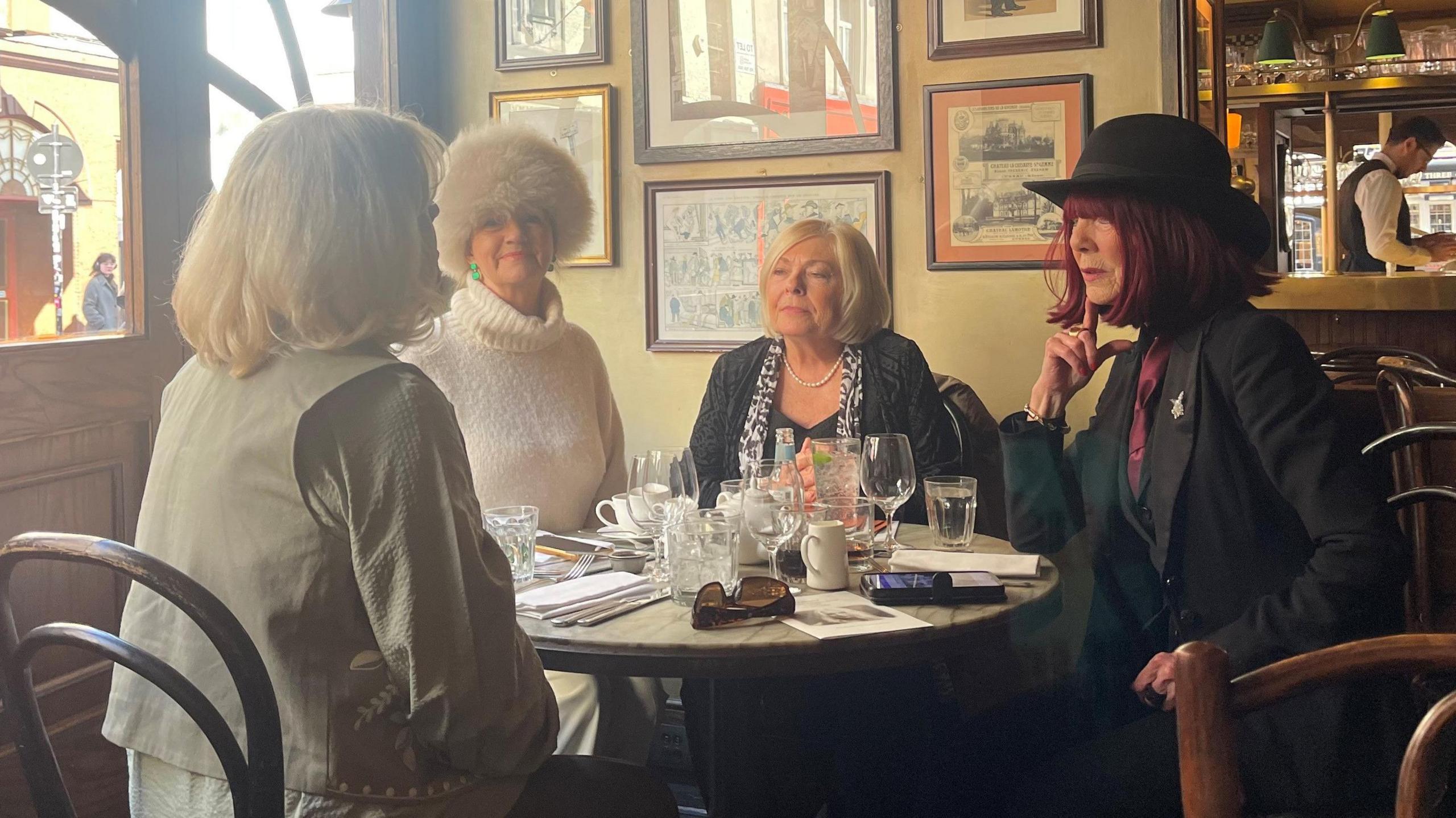 Four mature women sitting around a pub table near a window. All are dressed smartly, with one woman wearing a large cream furry hat and another a black trilby. They are chatting to each other and the table is laid with glasses and silverware. A waistcoat-wearing barman can be seen in the background polishing a glass.