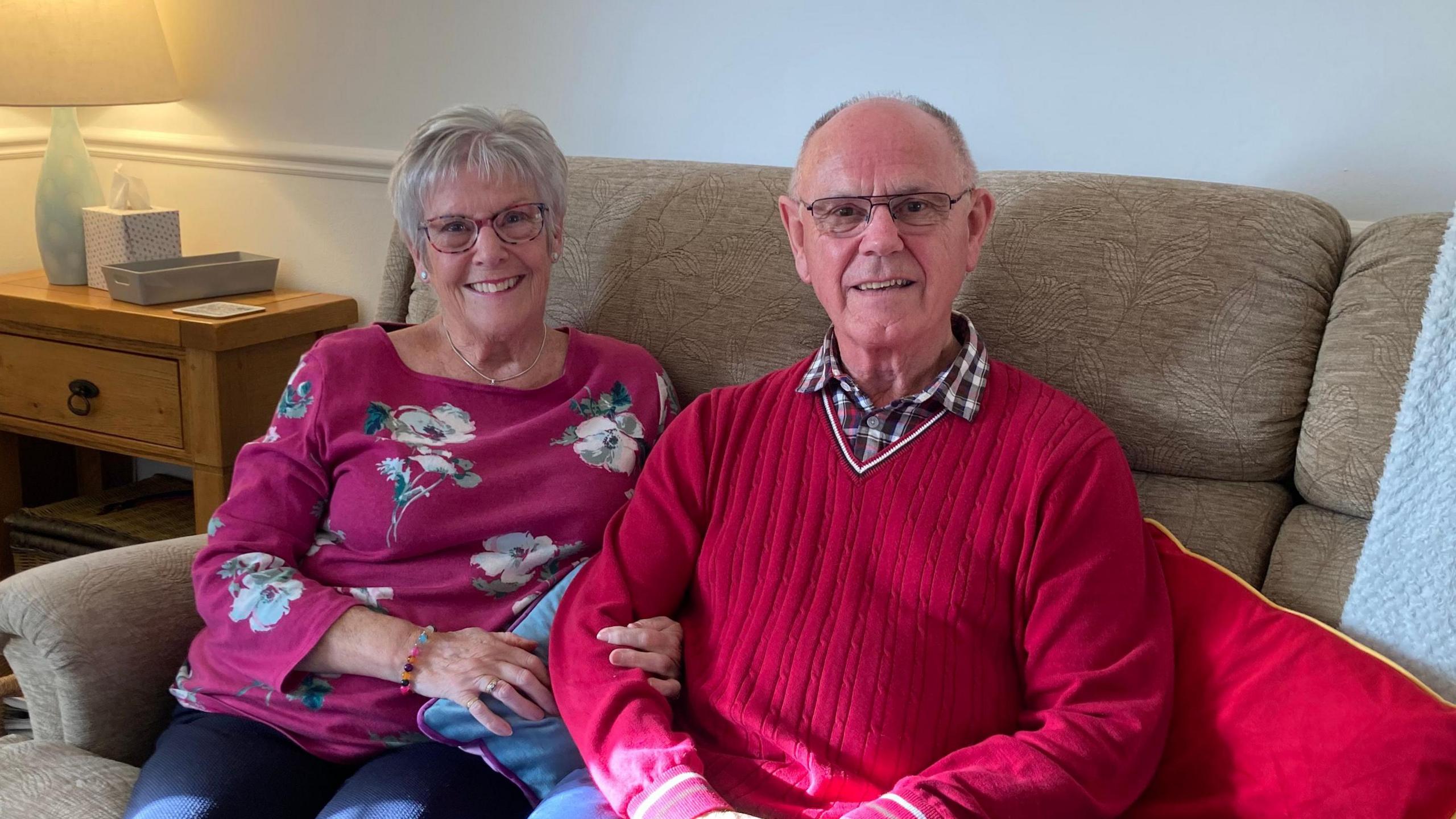 Karen and Colin Langlois are sitting on a sofa and smiling. Karan is wearing a pink top with flowers on it, and Colin is wearing a red jumper. Both are wearing spectacles.