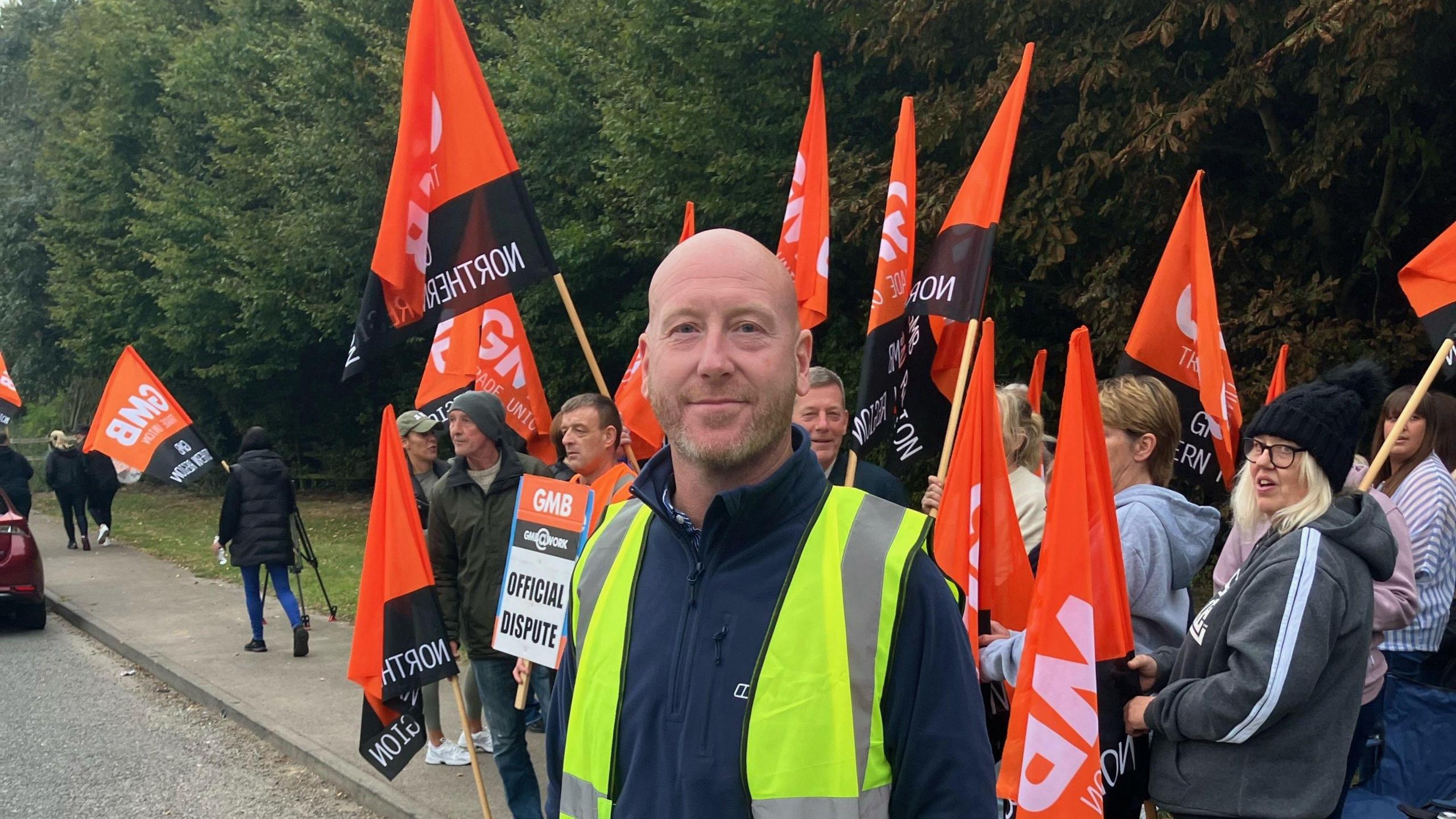 Dozens of picketers are waving flags in the background whilst a man, with a beard, looks into the camera. he is wearing a yellow high-vis jacket. The flags behind him are coloured orange and black, with "GMB" branding on them.