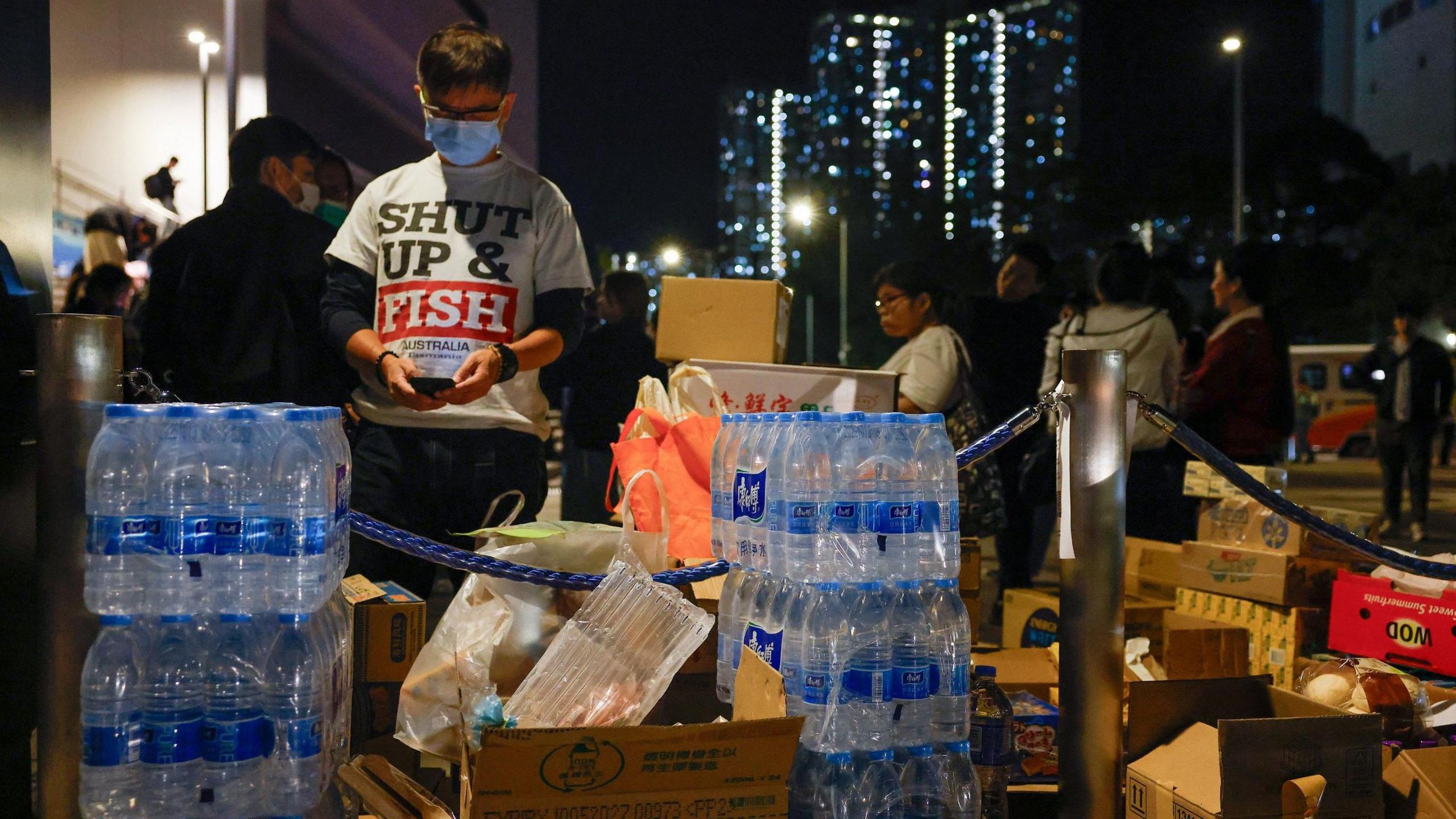 People standing next to aid supplies outdoors in the evening. There are cardboard boxes and water crates