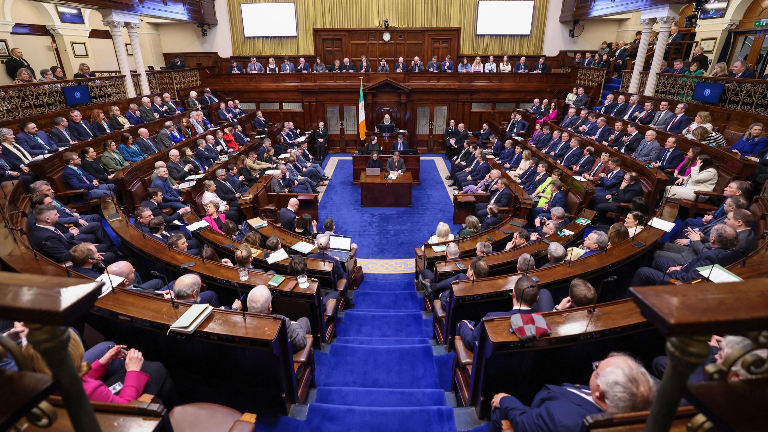 A full sitting of the Dail, the Irish Parliament from January 2025. The chamber has curved and straight wooden benches, a blue carpet and a podium in the back middle of the picture.