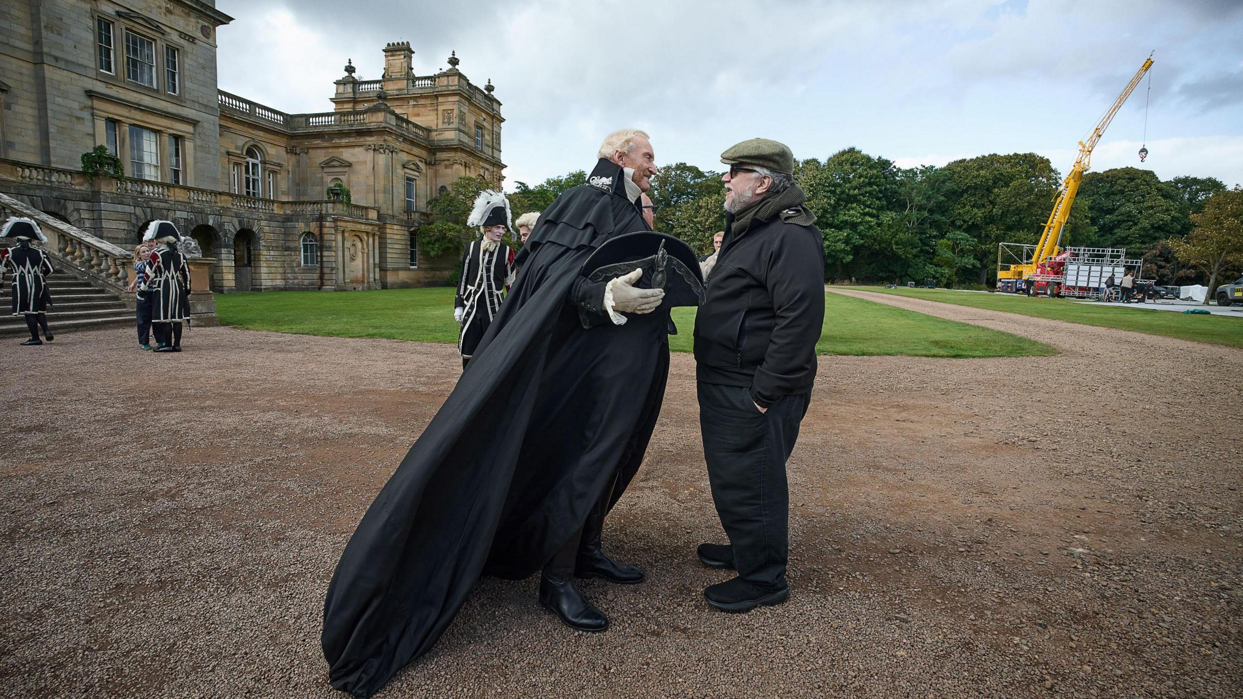 Another view of the outside of Gosford House exterior, with the actor Charles Dance - in flowing, black period cape and suit - speaking to Guillermo Del Toro, who is wearing a flat cap, black trousers and outdoor jacket. There is a cherry-picker care in the background and some extras in period costume. 
