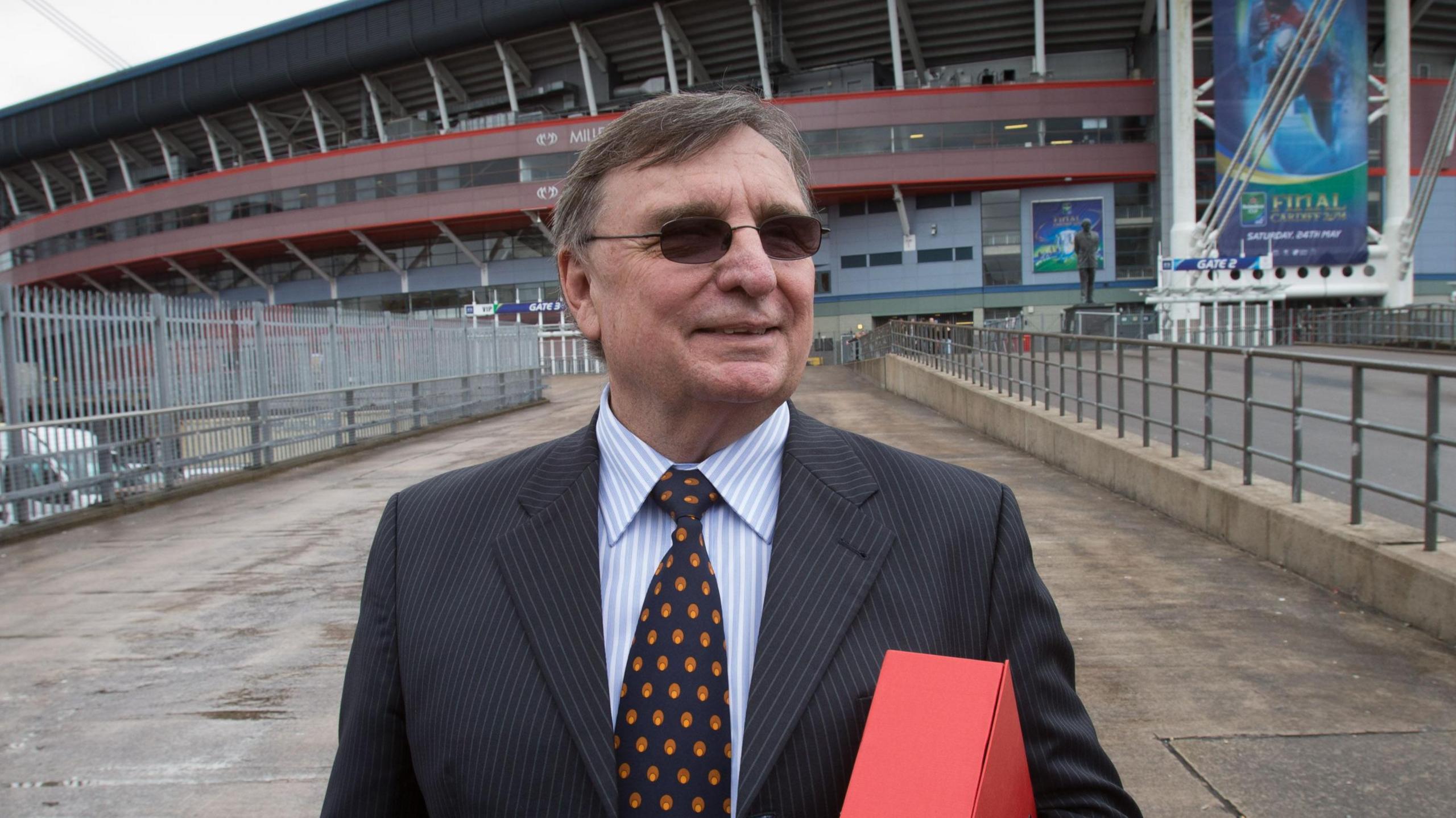 David Moffett wears a dark pin-striped suit, blue and red tie and dark glasses while holding a red box and standing in front of the Principality Stadium