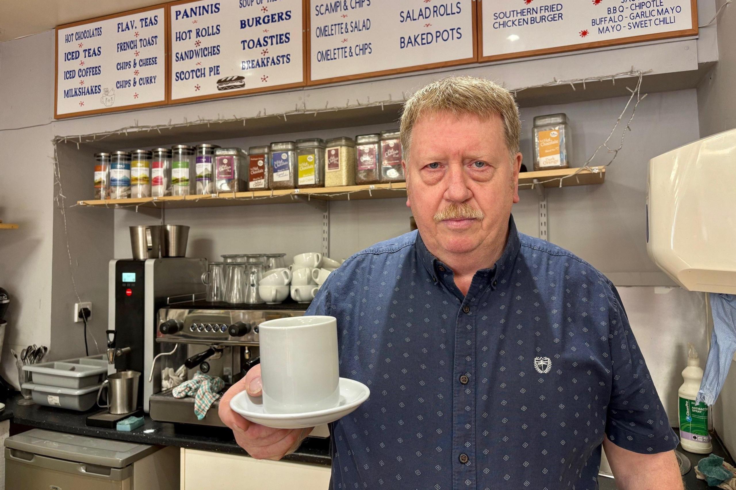 Jim is holding a coffee mug and saucer. He is wearing a blue shirt. There is a coffee machine in the background.