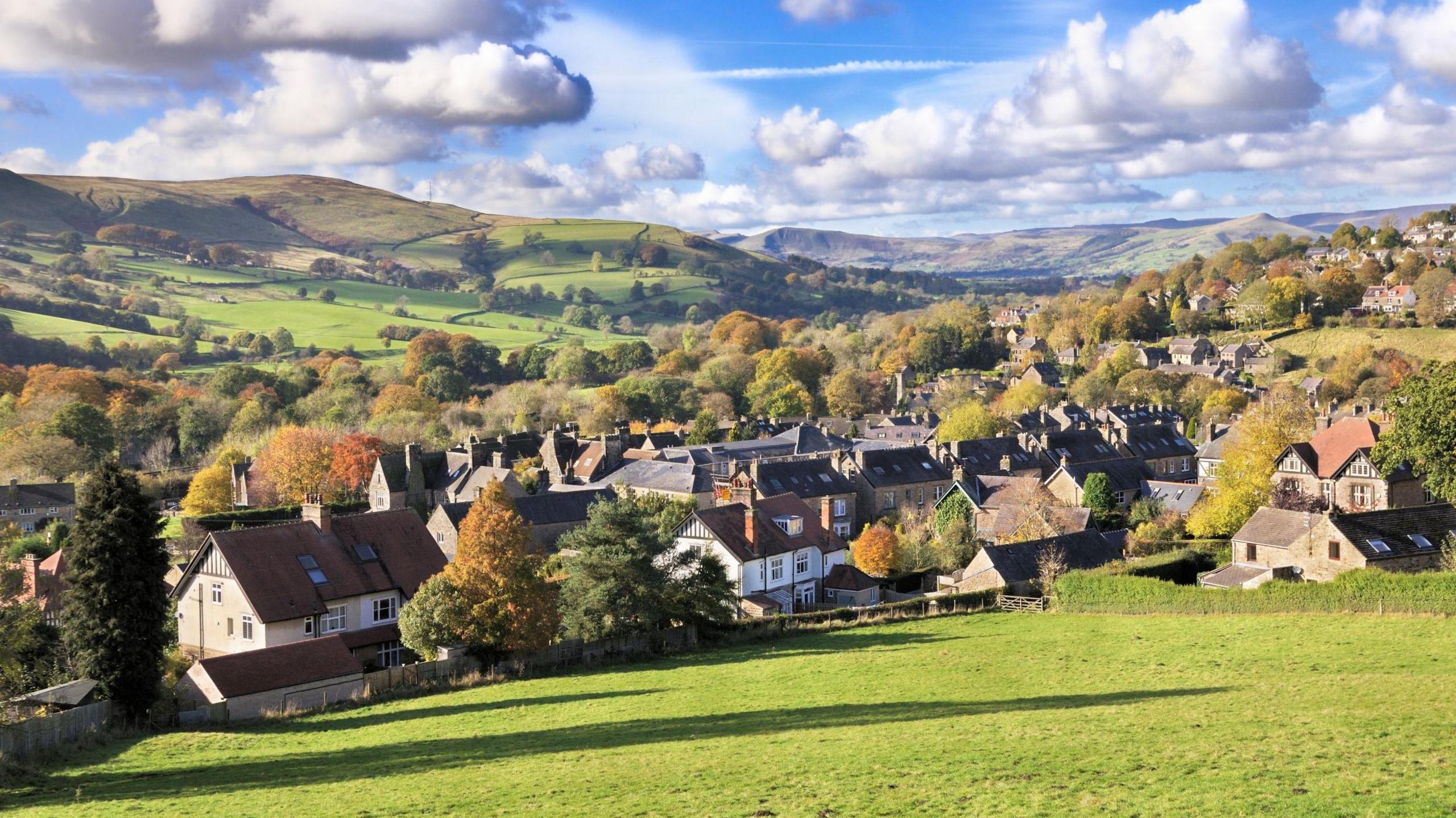 A photograph of the village of Hathersage. A number of houses can be seen surrounded by rolling hills and trees, and the sun is shining. 