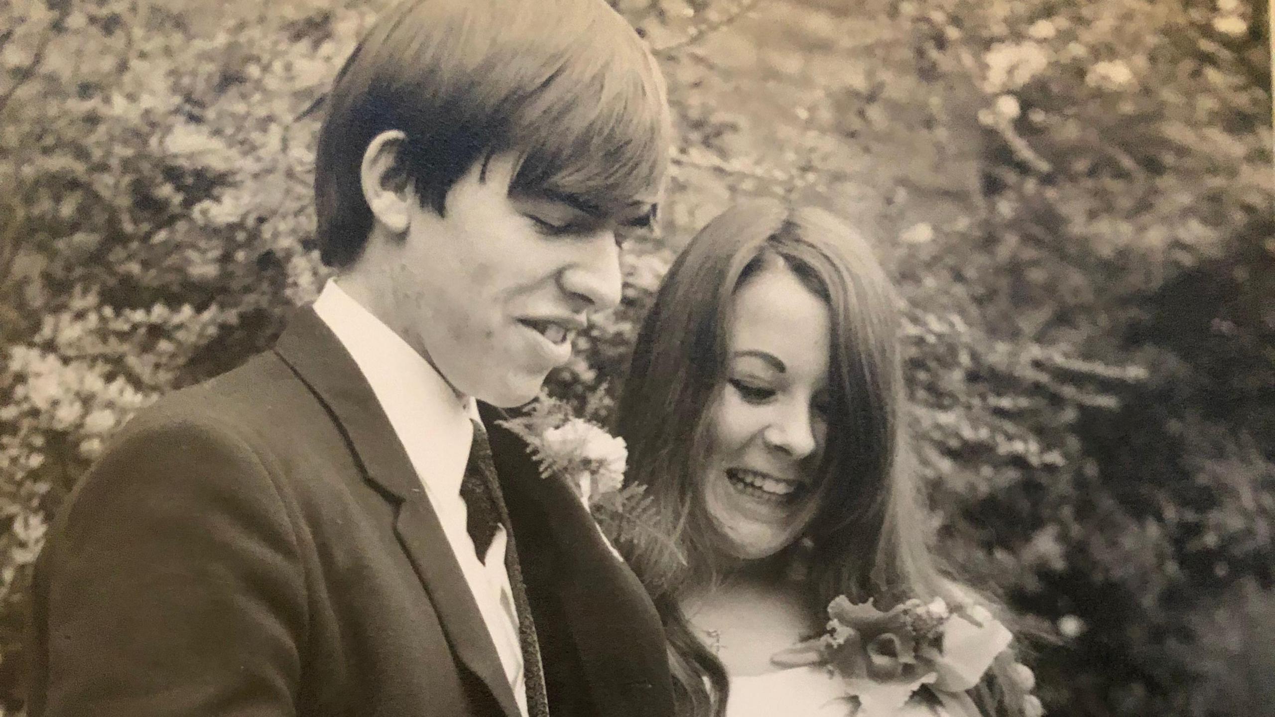 John wears a dark suit with a carnation buttonhole. Tina is wearing a white long-sleeved dress with a large floral buttonhole. They are smiling and looking down at their marriage certificate.