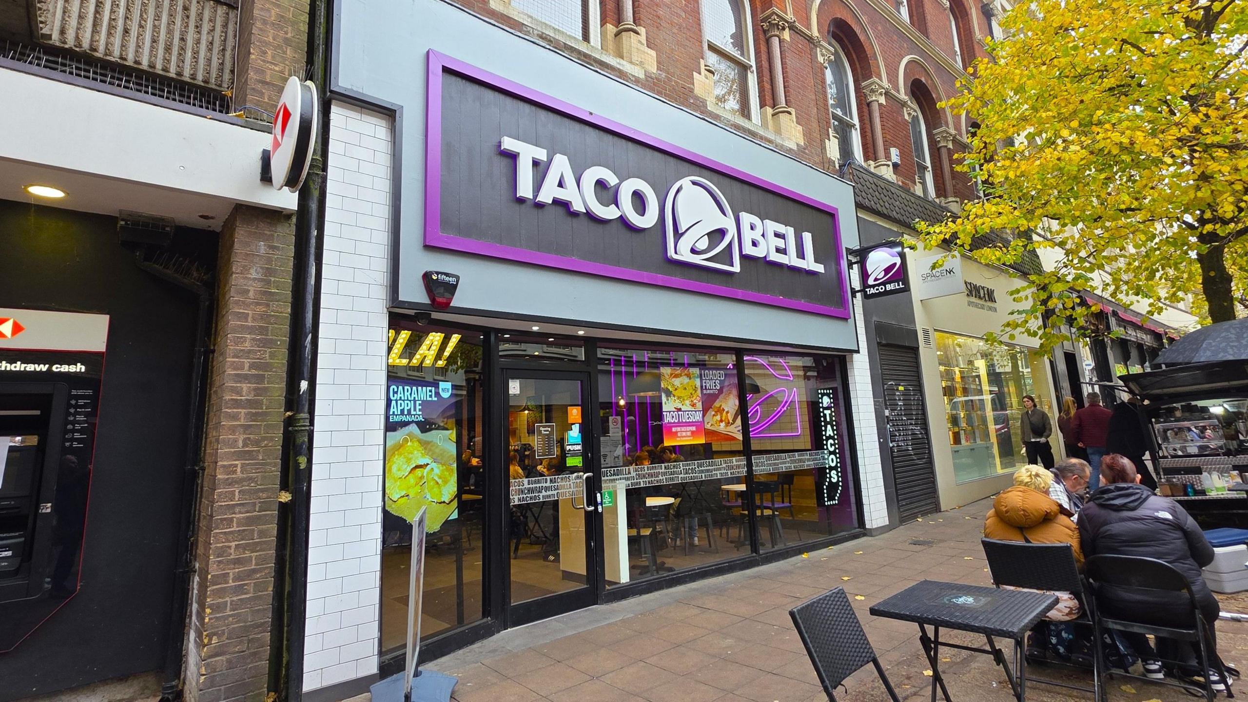The exterior of a Taco Bell. It's a small shopfront with a black and purple backing for the shop sign, which also features a bell. Outside we can see a couple of tables and chairs.