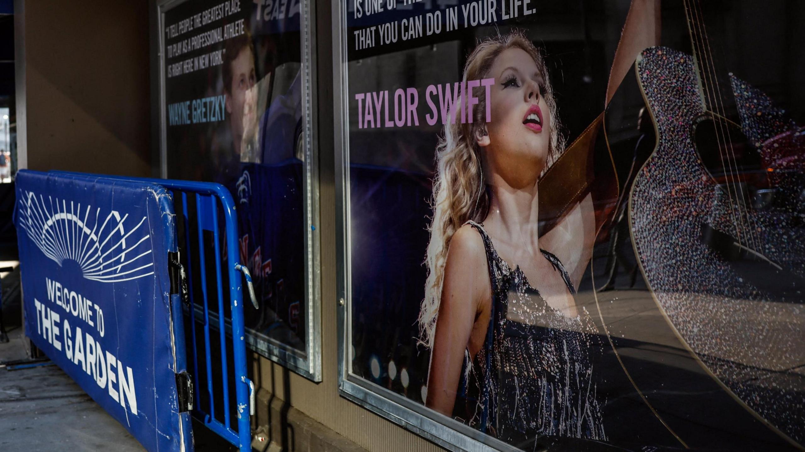 A poster of Taylor Swift  holding up a guitar outside Madison Square Garden, next to crush barriers saying 