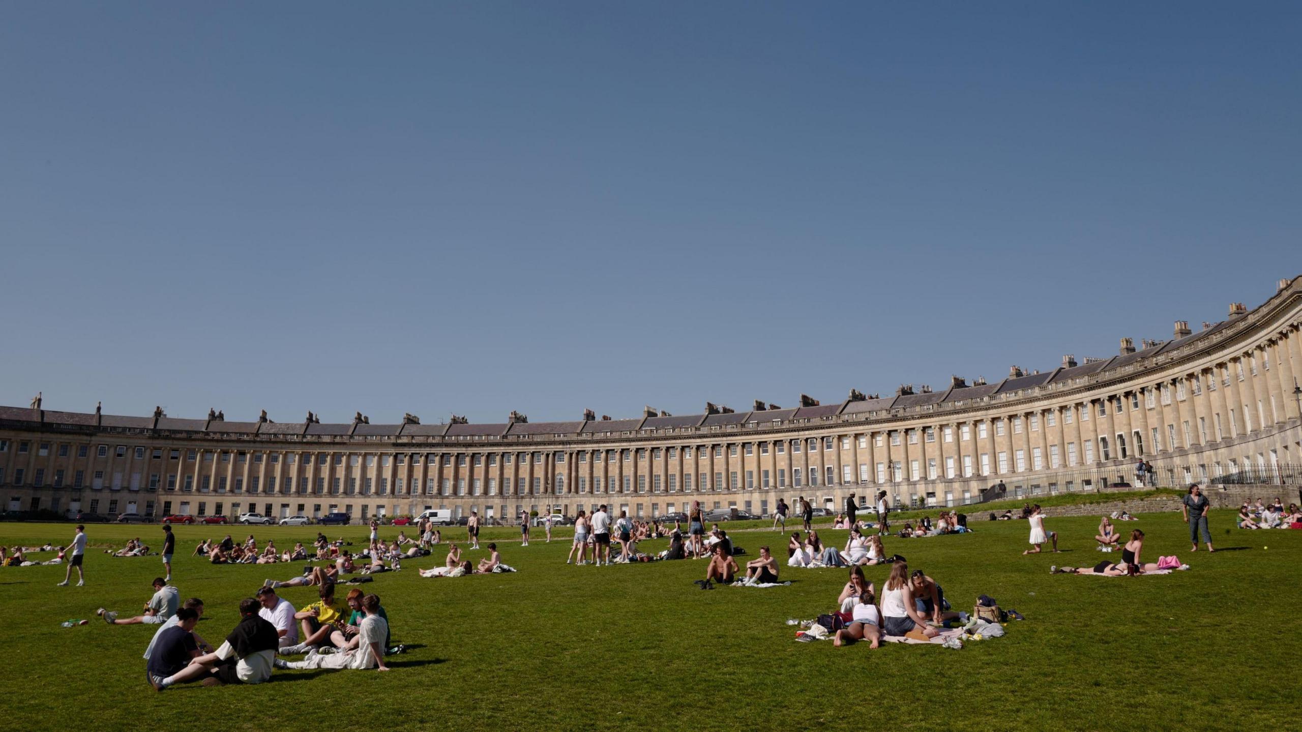 Groups of people sunbathe in the wide grass area in front of the Royal Crescent in Bath. The sky is clear and blue overhead