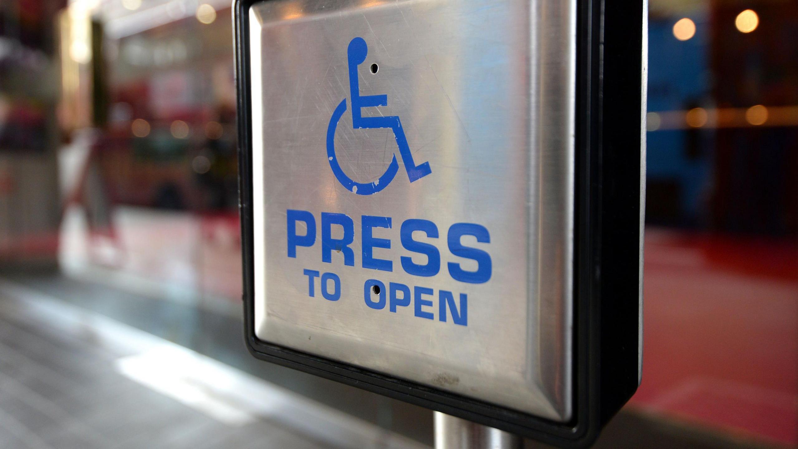 A square silver button used to open automatic doors. It features a blue line drawing of a stick person in a wheelchair and the words PRESS TO OPEN.