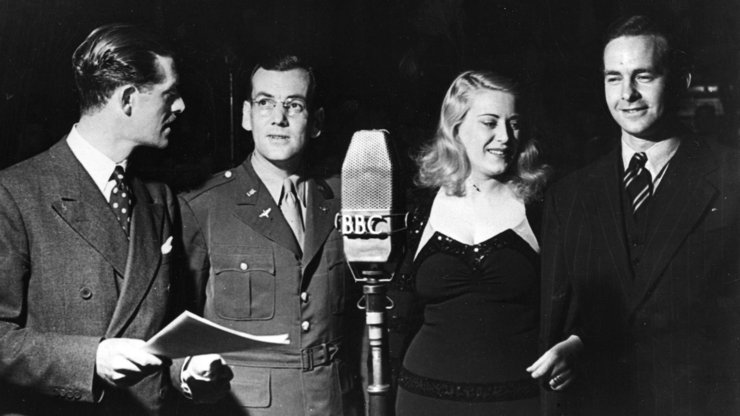 The four musicians stand around a large microphone with BBC labelled on the front. The photo is black and white. Glenn Miller, second from left, is wearing a military jacket over a shift and tie, and round glasses. He looks up at Leslie Mitchell who is holding a sheaf of papers. Mitchell and Trent wear a spotty and stripey tie respectively, while Dorothy wears a figure-hugging black dress and has blonde shoulder length wavy hair. 