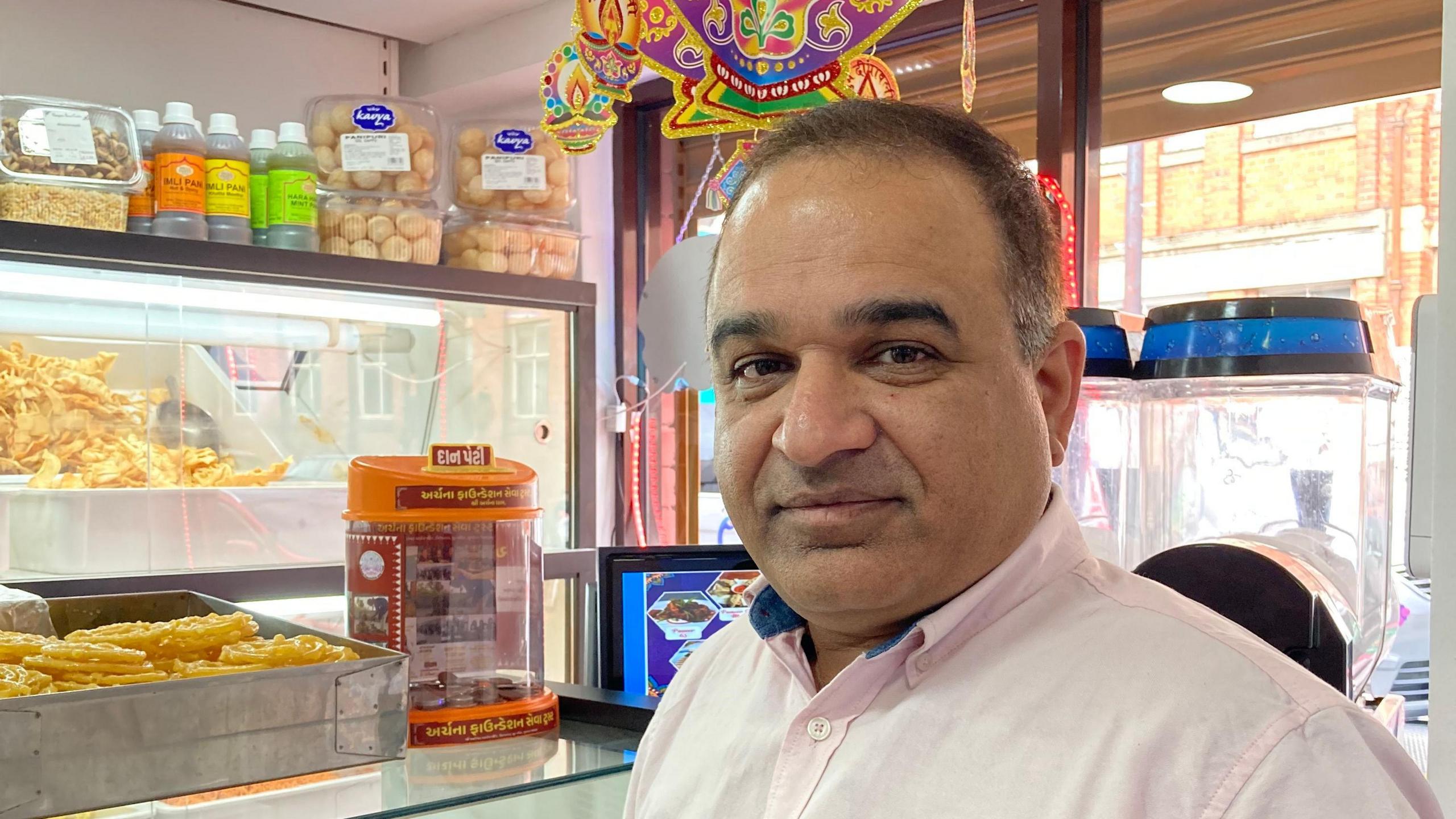 A man with short, dark hair, turning grey at the temples, looks into the camera. He wears a very light pink shirt. Behind him are shelves with bottles and jars of sweets.